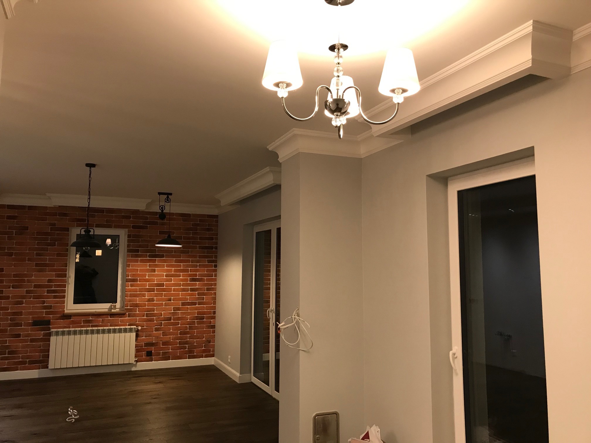 Wykończone wnętrze: ściana z cegły, drewniana podłoga, szare ściany, białe listwy przypodłogowe i sufitowe, ozdobna lampa sufitowa, wiszące lampy z czarnymi kloszami, biały grzejnik pod oknem.