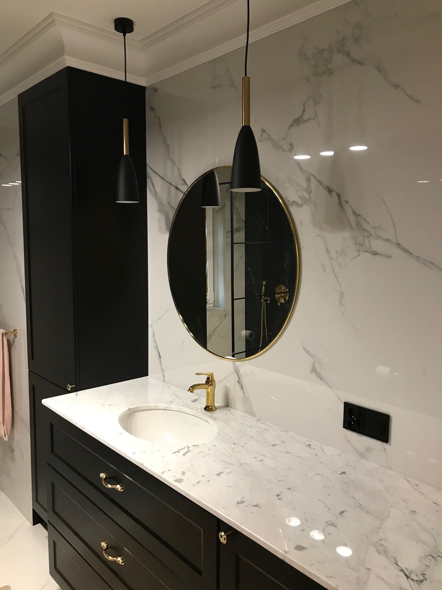 Elegancka łazienka z czarnymi szafkami, marmurowym blatem i ścianami, okrągłym lustrem w złotej ramie oraz złotą armaturą, oświetlona wiszącymi lampami z elementami w kolorze złota.