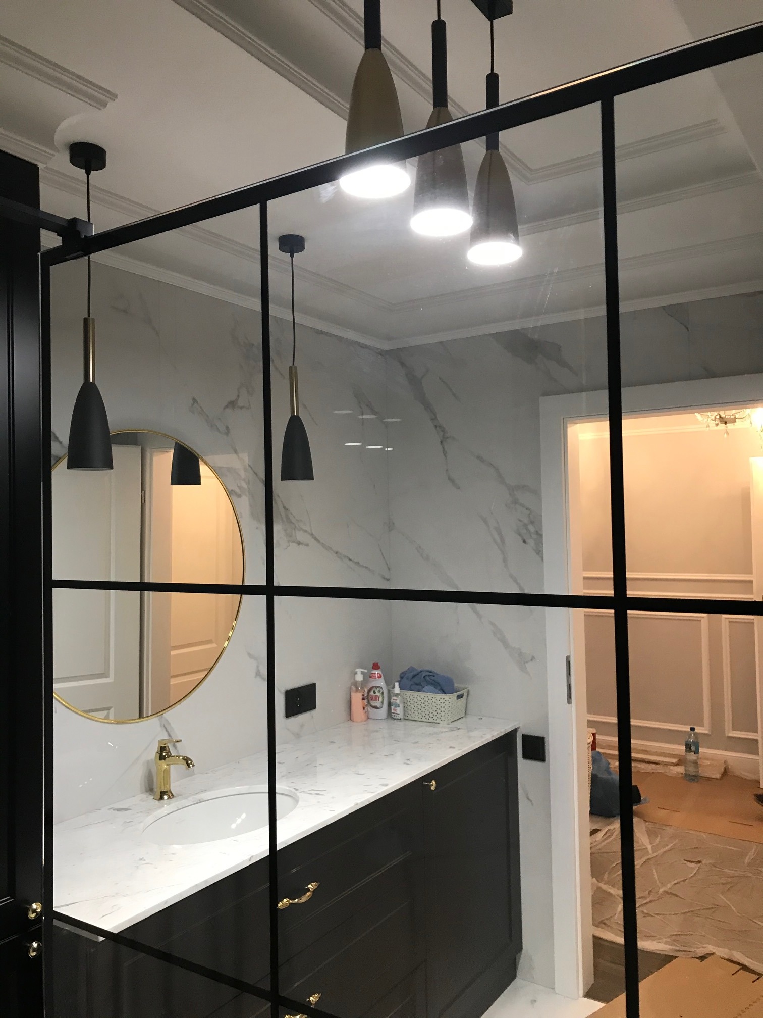 Nowoczesna łazienka z czarną ramą szklanej kabiny prysznicowej, marmurową ścianą, złotą armaturą i minimalistycznymi lampami wiszącymi.