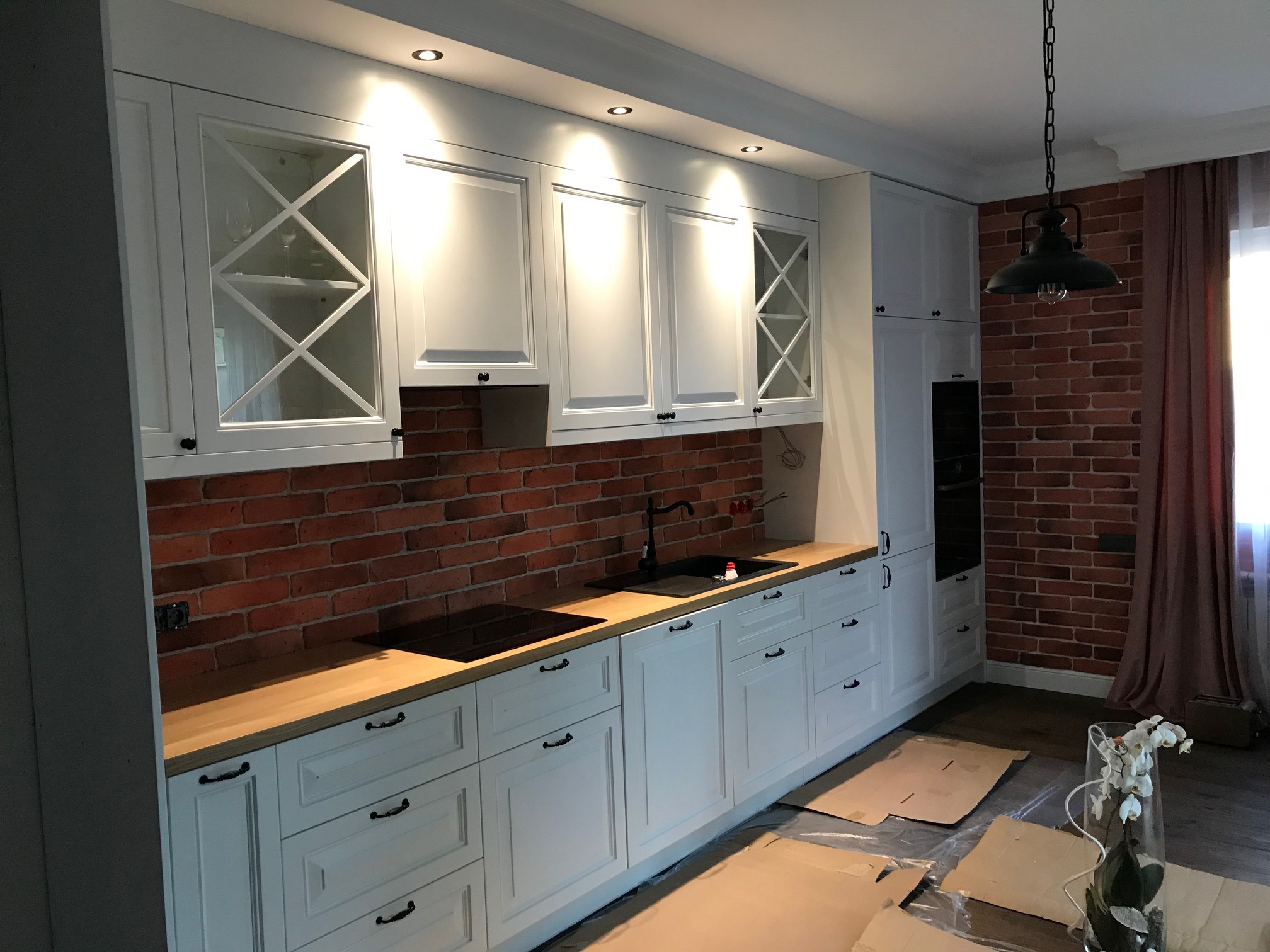 Jasna kuchnia w stylu rustykalnym z białymi szafkami, drewnianym blatem i ścianą z czerwonej cegły, oświetlona punktowymi lampami sufitowymi i wiszącą lampą industrialną, z widocznymi kartonami...