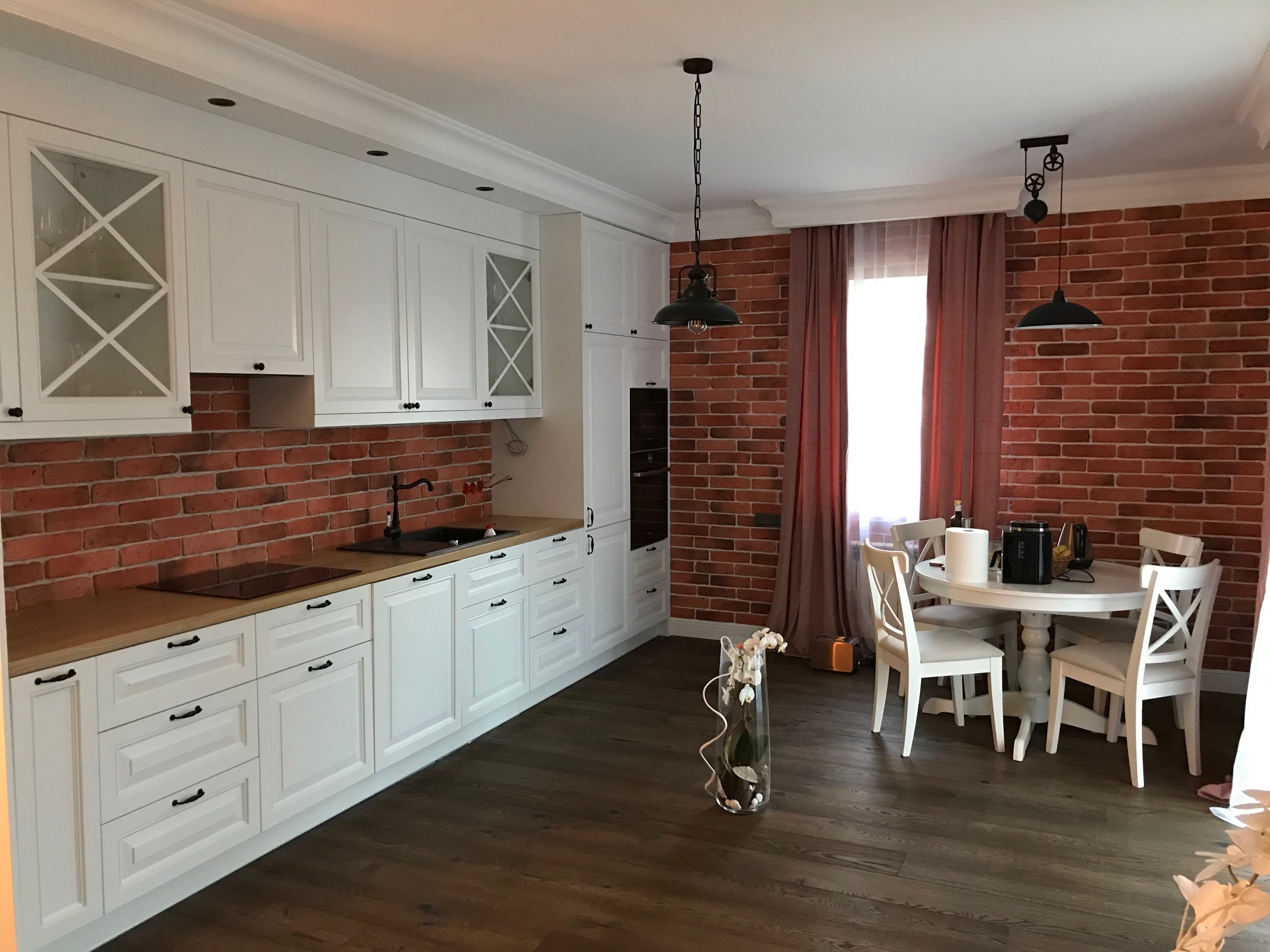 Jasna kuchnia z białymi szafkami, drewnianym blatem i ceglaną ścianą, uzupełniona czarną armaturą i lampami w stylu industrialnym, z widocznym stołem jadalnym na drewnianej podłodze.