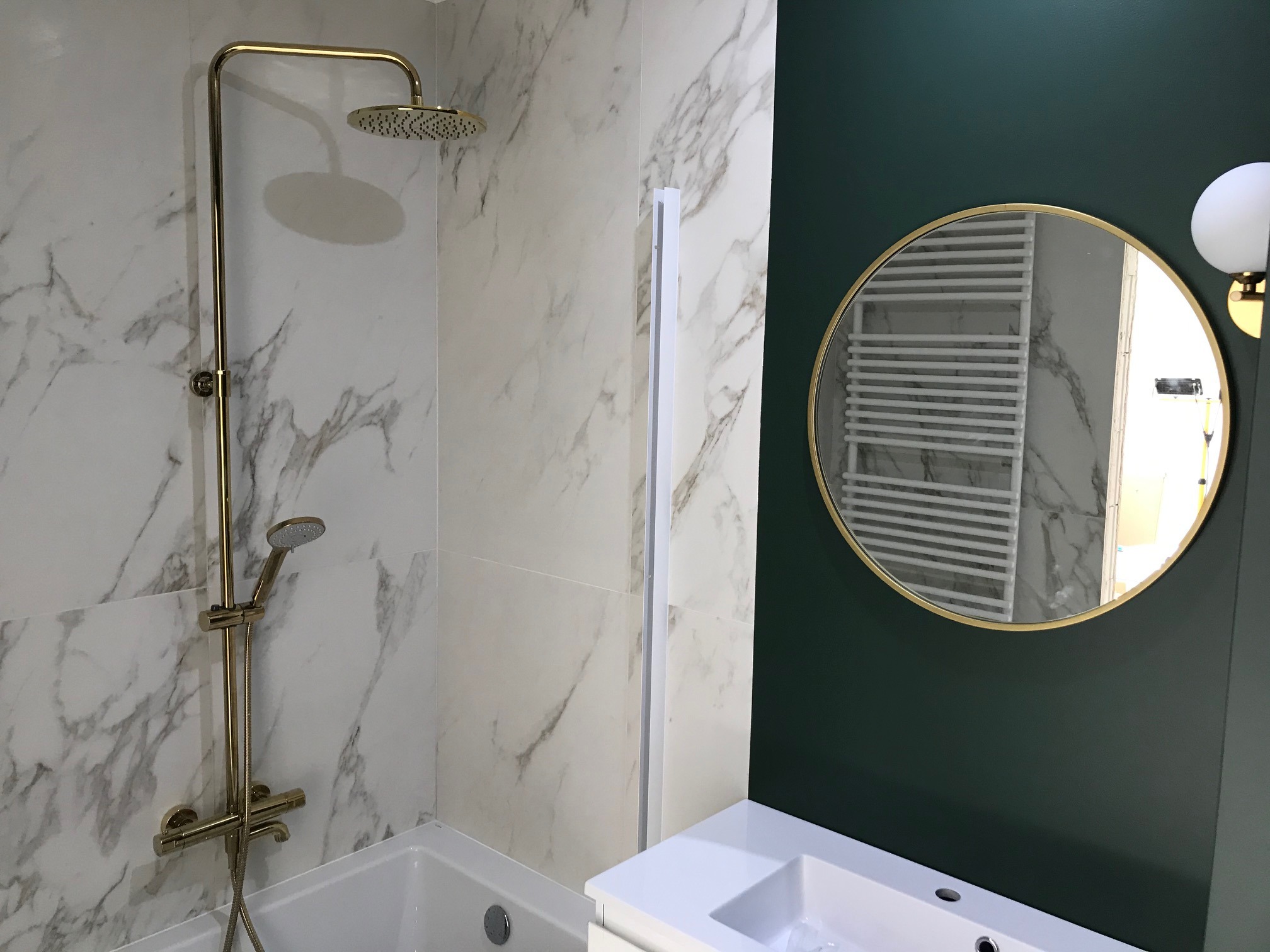 Nowoczesna łazienka z marmurowymi płytkami, złotą armaturą prysznicową i okrągłym lustrem na ciemnozielonej ścianie.