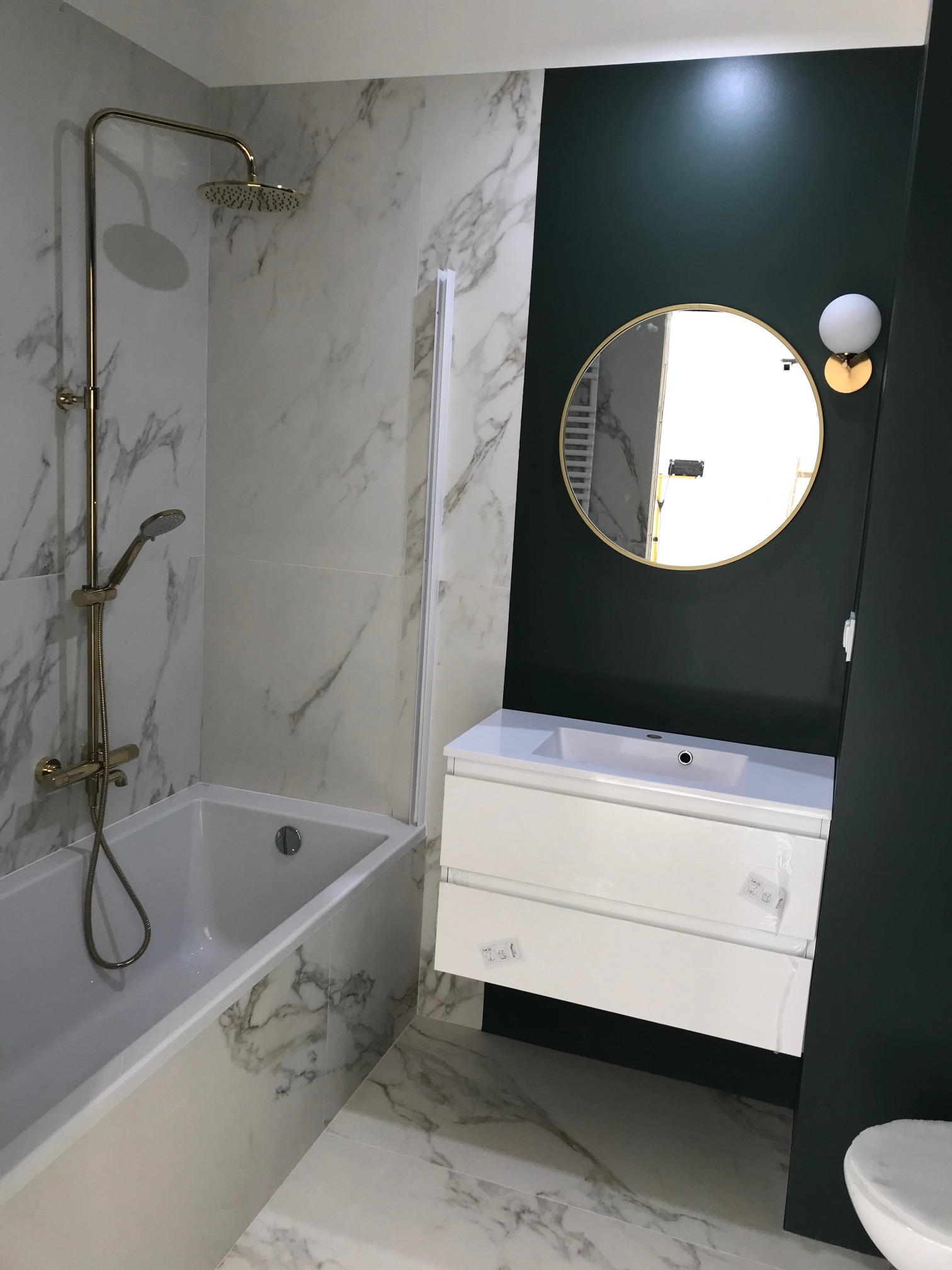 Nowoczesna łazienka z wanną obudowaną płytkami imitującymi marmur, złotą armaturą, umywalką z szafką w kolorze białym, okrągłym lustrem w złotej ramie i ciemnozieloną ścianą.
