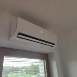 Biały klimatyzator marki Rotenso zamontowany na ścianie nad oknem z widokiem na drzewa i dom, wyświetlający temperaturę 22 stopnie.