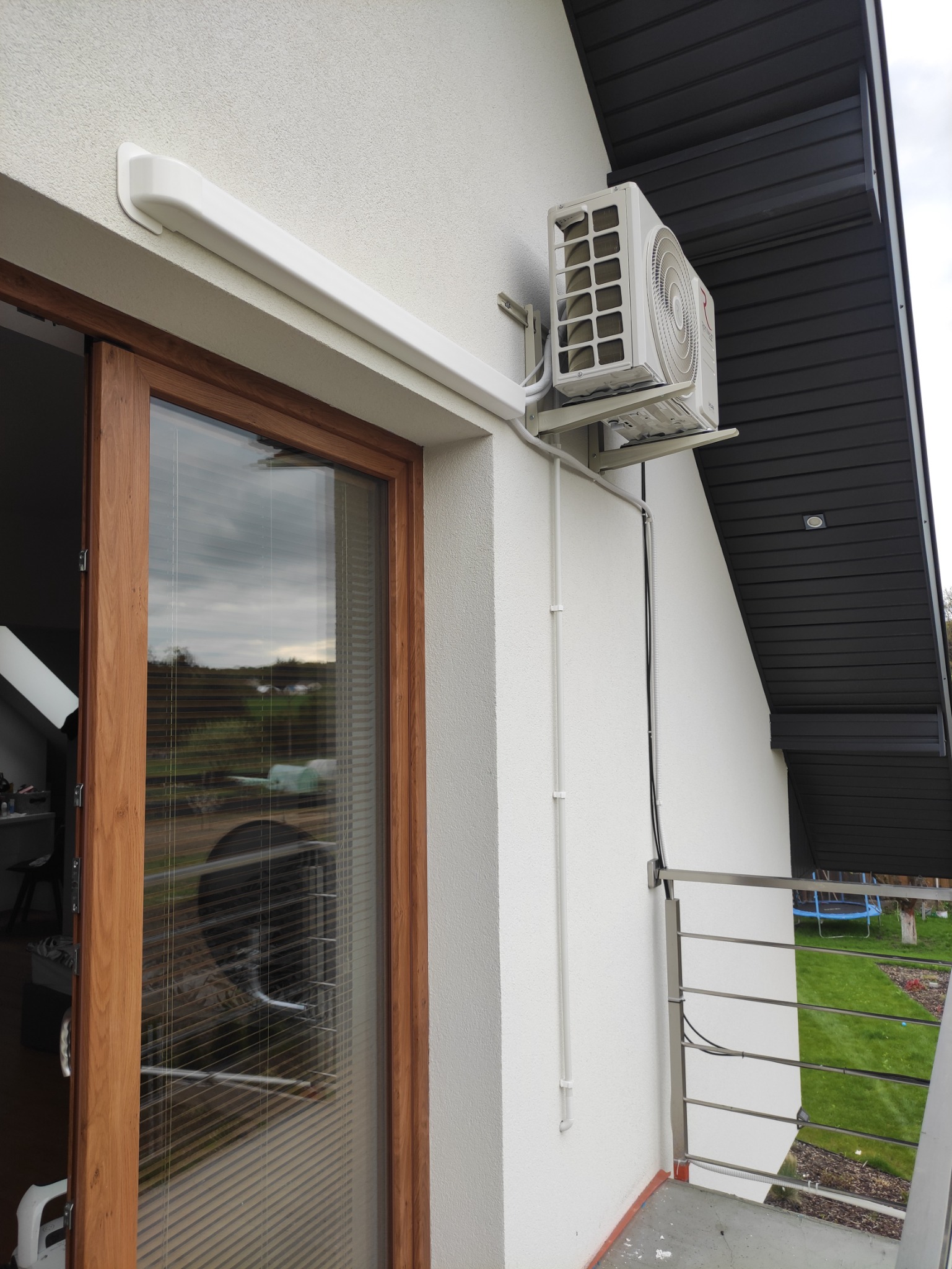 Zewnętrzna jednostka klimatyzacji zamontowana na białej elewacji budynku w Puławach, widoczne przewody i rury prowadzące do wnętrza przez okno balkonowe.