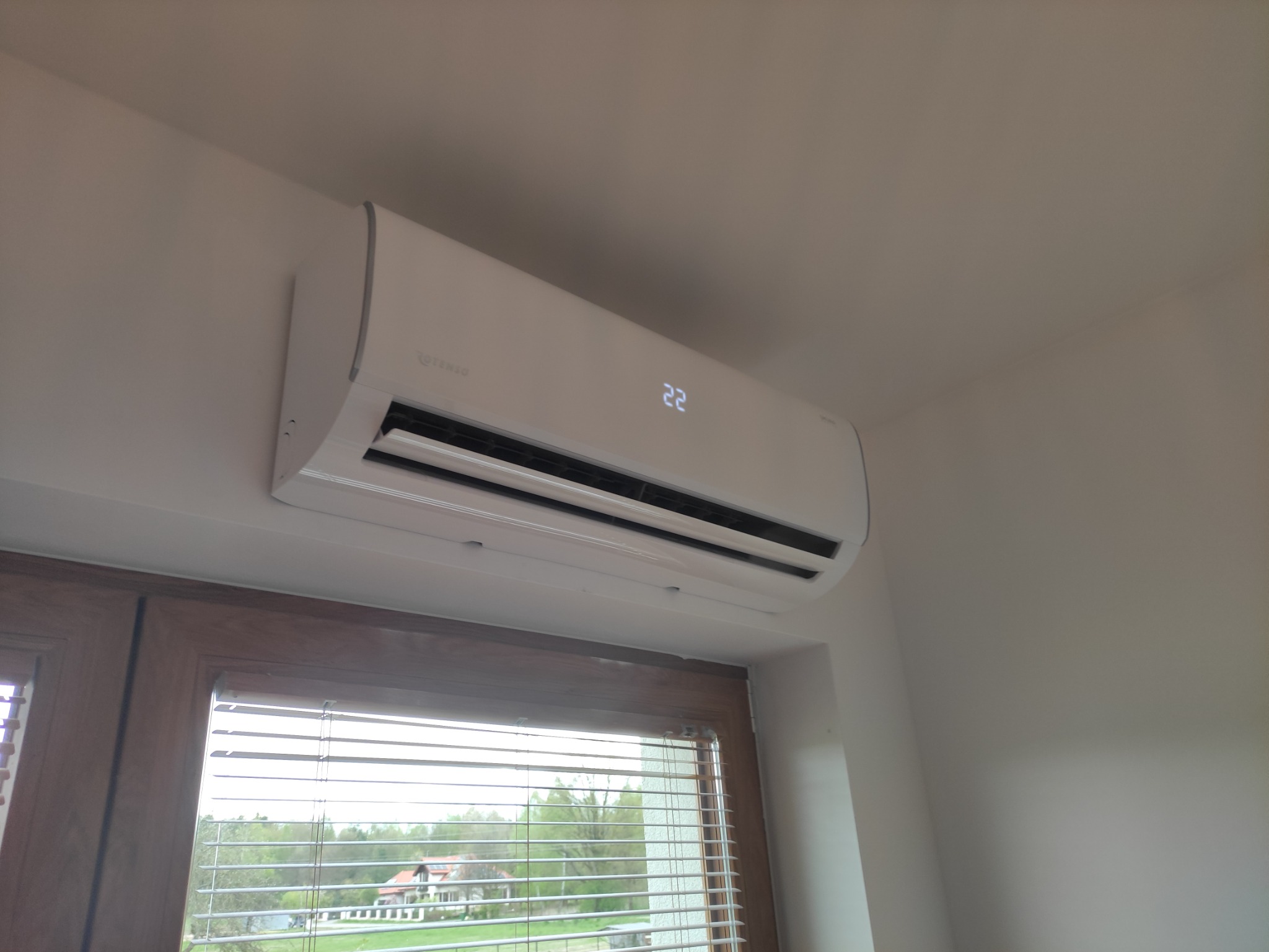 Biały klimatyzator marki Rotenso zamontowany na ścianie nad oknem z widokiem na drzewa i dom, wyświetlający temperaturę 22 stopnie.