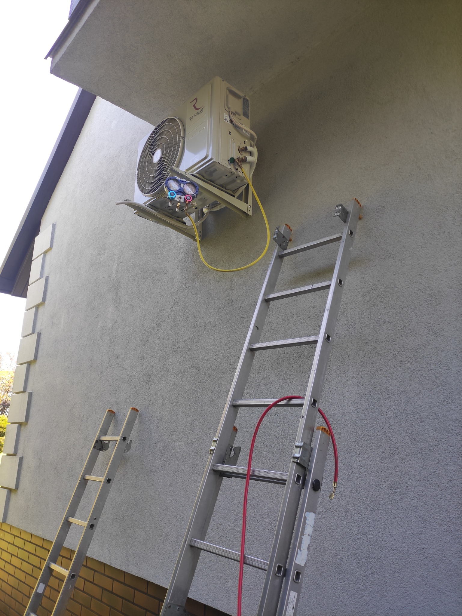 Zewnętrzna jednostka klimatyzacji Rotenso zamontowana na szarej elewacji budynku, widoczna z poziomu dwóch rozstawionych drabin, z podłączonymi przewodami pomiarowymi.