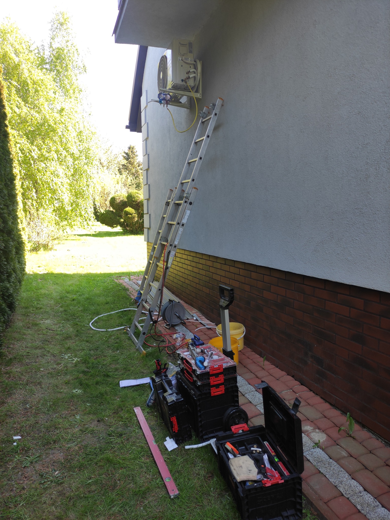 Zewnętrzna jednostka klimatyzacji zamontowana na elewacji budynku, z podłączonymi przewodami i manometrami; rozstawiona drabina i rozłożone skrzynki z narzędziami na trawie.