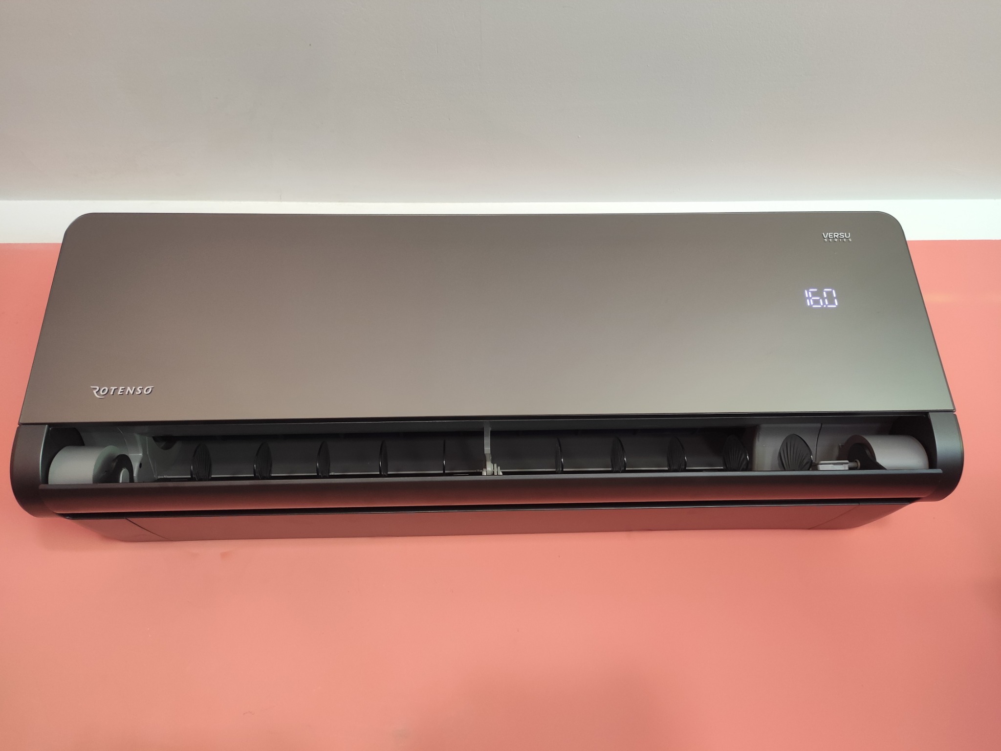 Szary klimatyzator Rotenso Versu Series zamontowany na różowej ścianie, wyświetlający temperaturę 16 stopni.