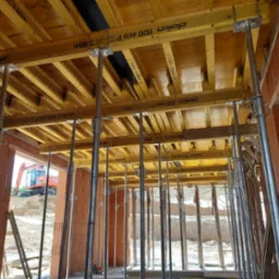 Wnętrze w trakcie budowy domu murowanego z widocznym szalunkiem stropu Doka H20 eco P podpartym stalowymi stemplami, w tle wykop i koparka.