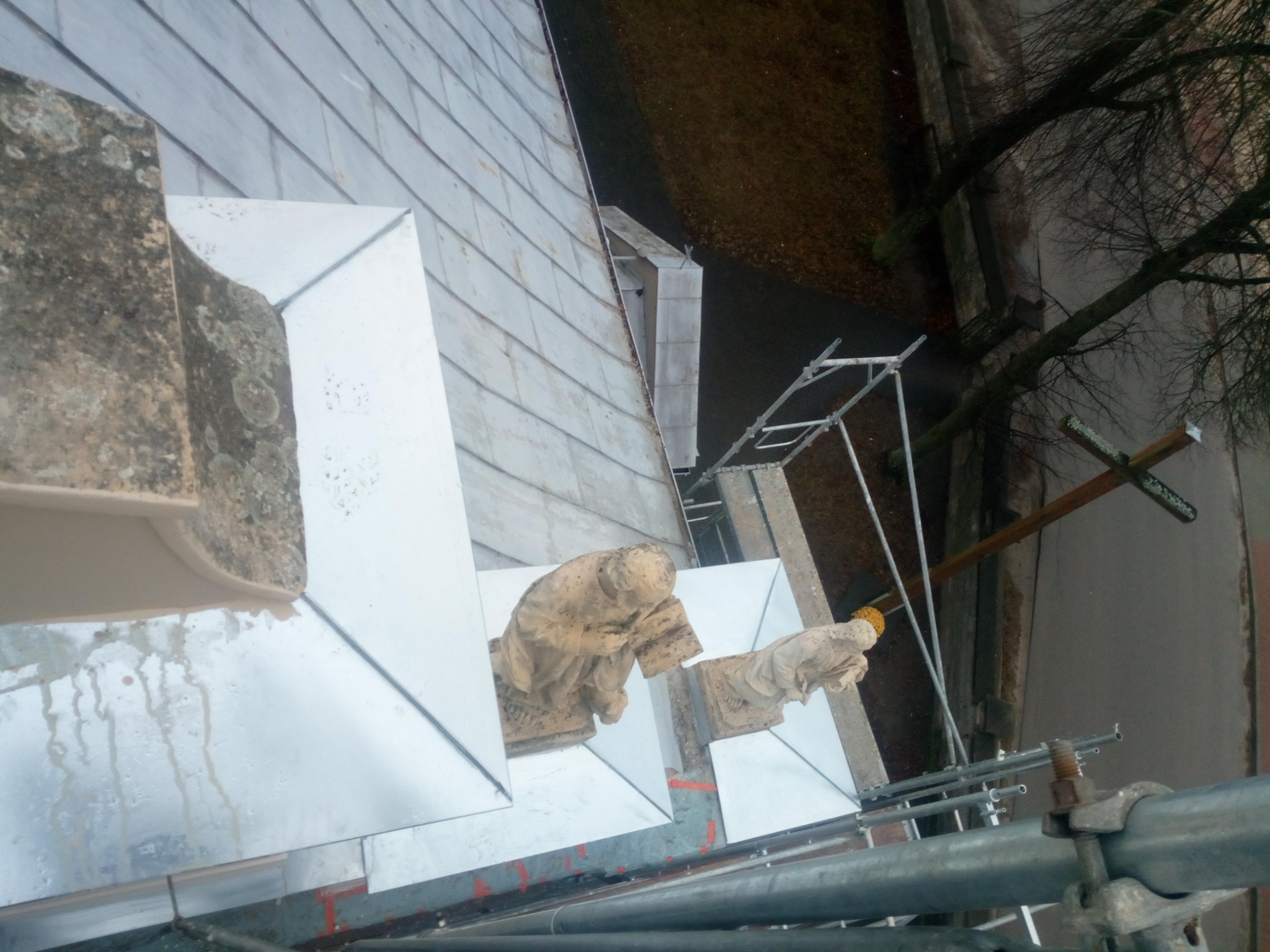 Widok z góry na fragment dachu z nowymi, błyszczącymi obróbkami blacharskimi wokół kamiennych elementów dekoracyjnych; widoczne rusztowanie i fragment otoczenia.