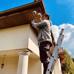 Adius sp. z o.o. - Instalator na drabinie montuje kamerę kopułkową pod brązowym okapem domu z beżową elewacją, widok z dołu na błękitne niebo z chmurami.
