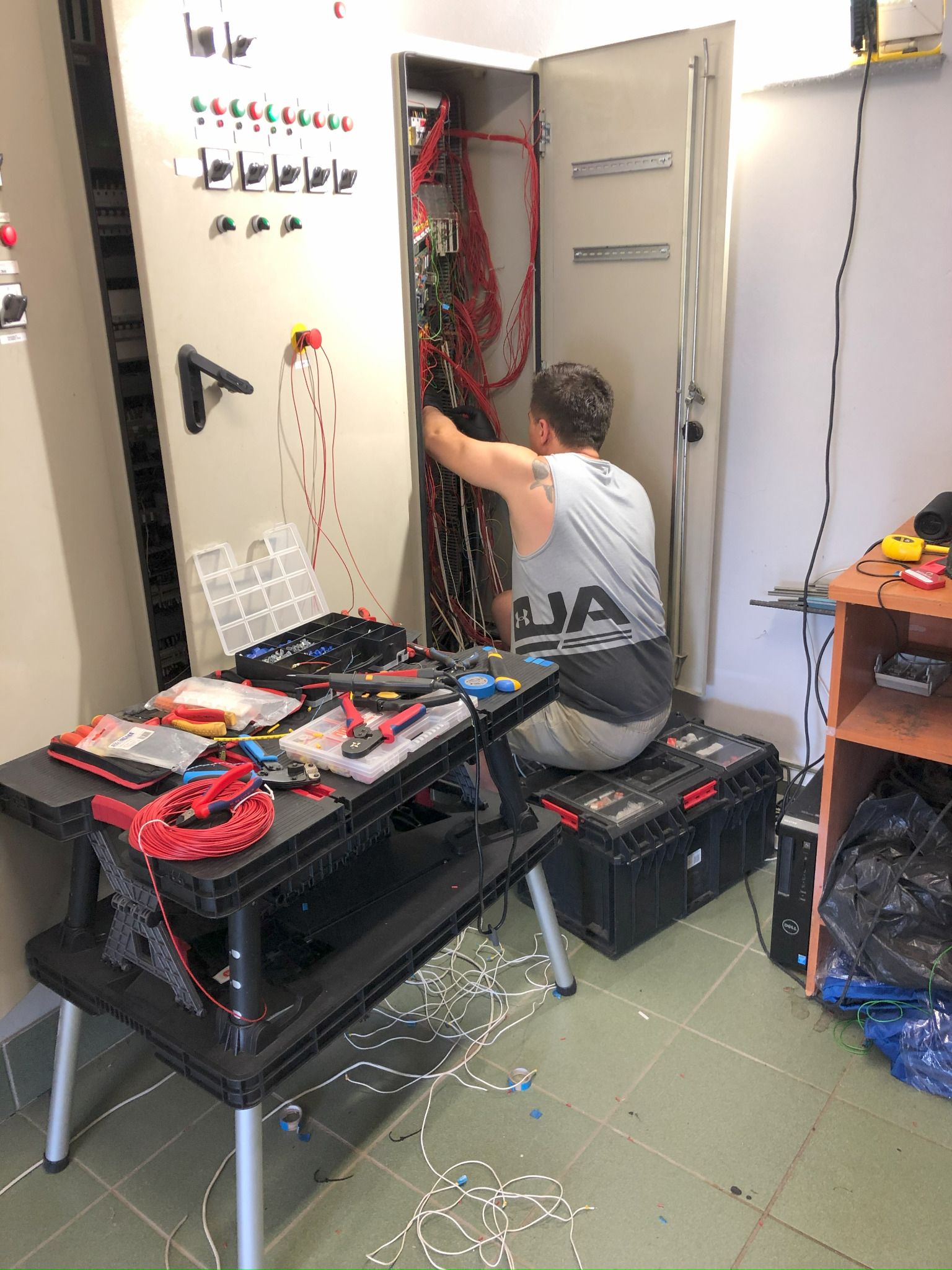 Elektryk w szarej koszulce z tatuażem na ramieniu pracuje nad okablowaniem wewnątrz otwartej szafy sterowniczej, siedząc na skrzynkach narzędziowych, z rozłożonymi narzędziami i przewodami...
