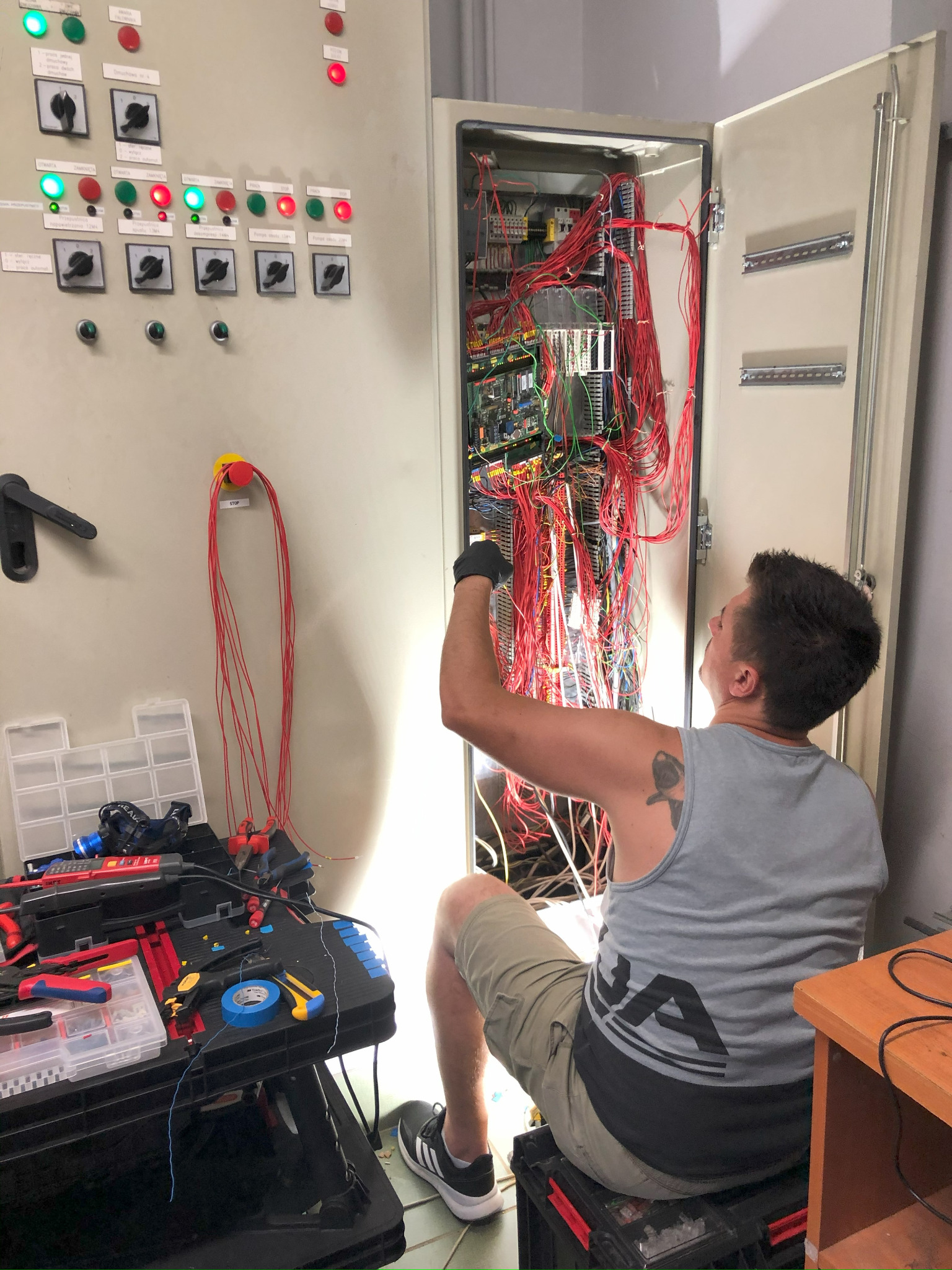 Elektryk w szarym podkoszulku z tatuażem na ramieniu, siedząc na skrzynce narzędziowej, pracuje przy otwartej szafie sterowniczej z gąszczem czerwonych kabli. Na pierwszym planie widoczny stół...