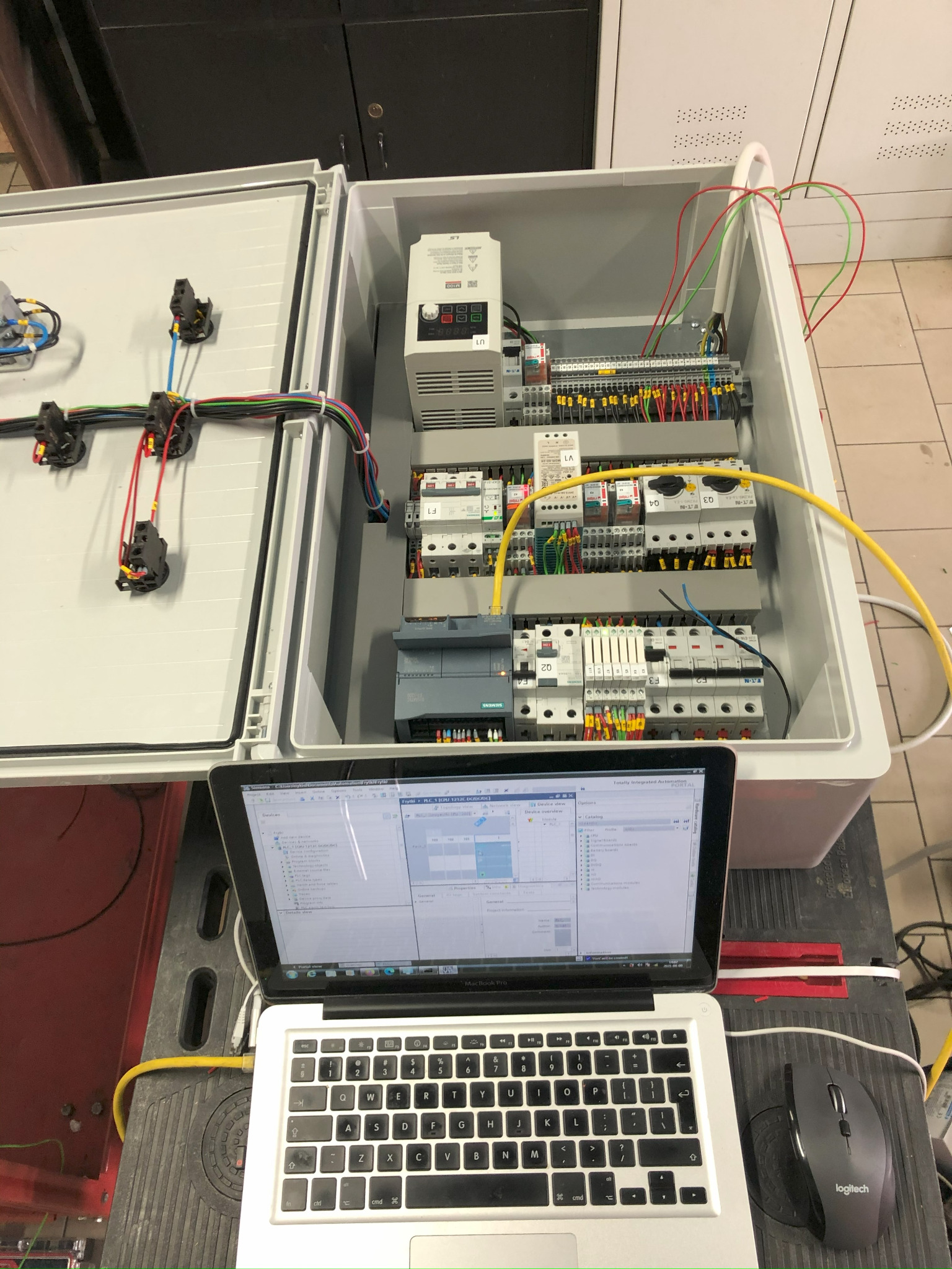 Otwarta szafa sterownicza z zainstalowanymi modułami, przewodami i podłączonym laptopem z uruchomionym oprogramowaniem do konfiguracji sterownika PLC Siemens, widok z góry.