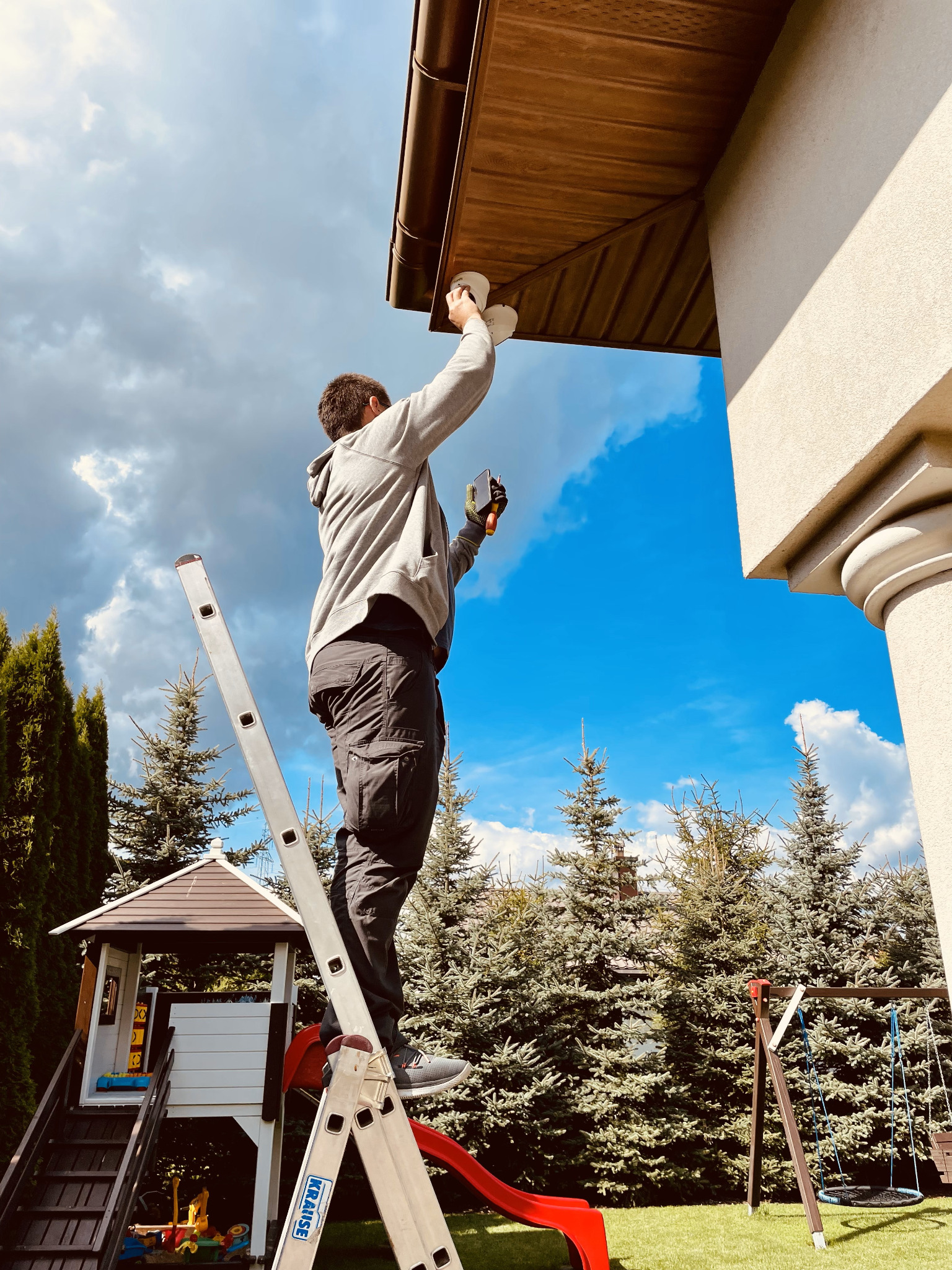Instalator na drabinie montuje białą lampę pod okapem domu, widok z dołu na tle błękitnego nieba i zielonych drzew.