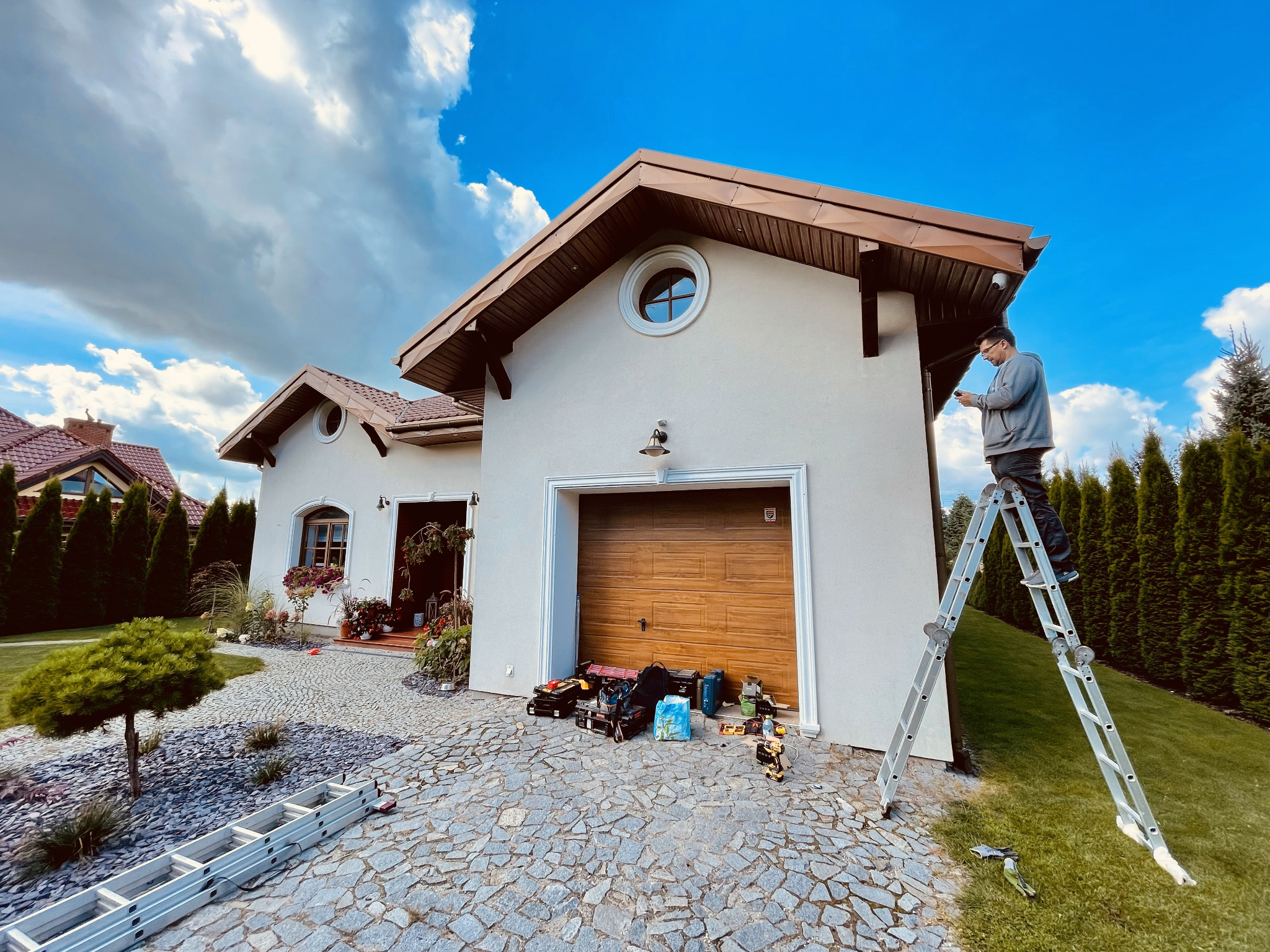 Instalator na drabinie przy domu z narzędziami, prawdopodobnie montujący kamerę monitoringu pod okapem dachu. Widoczne skrzynki narzędziowe i akcesoria na brukowanym podjeździe.