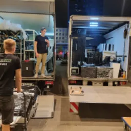 Dwa busy z otwartymi przestrzeniami ładunkowymi podczas załadunku, widoczni pracownicy w trakcie przenoszenia wyposażenia, jeden z wózkiem, drugi z kubkiem w ręku.