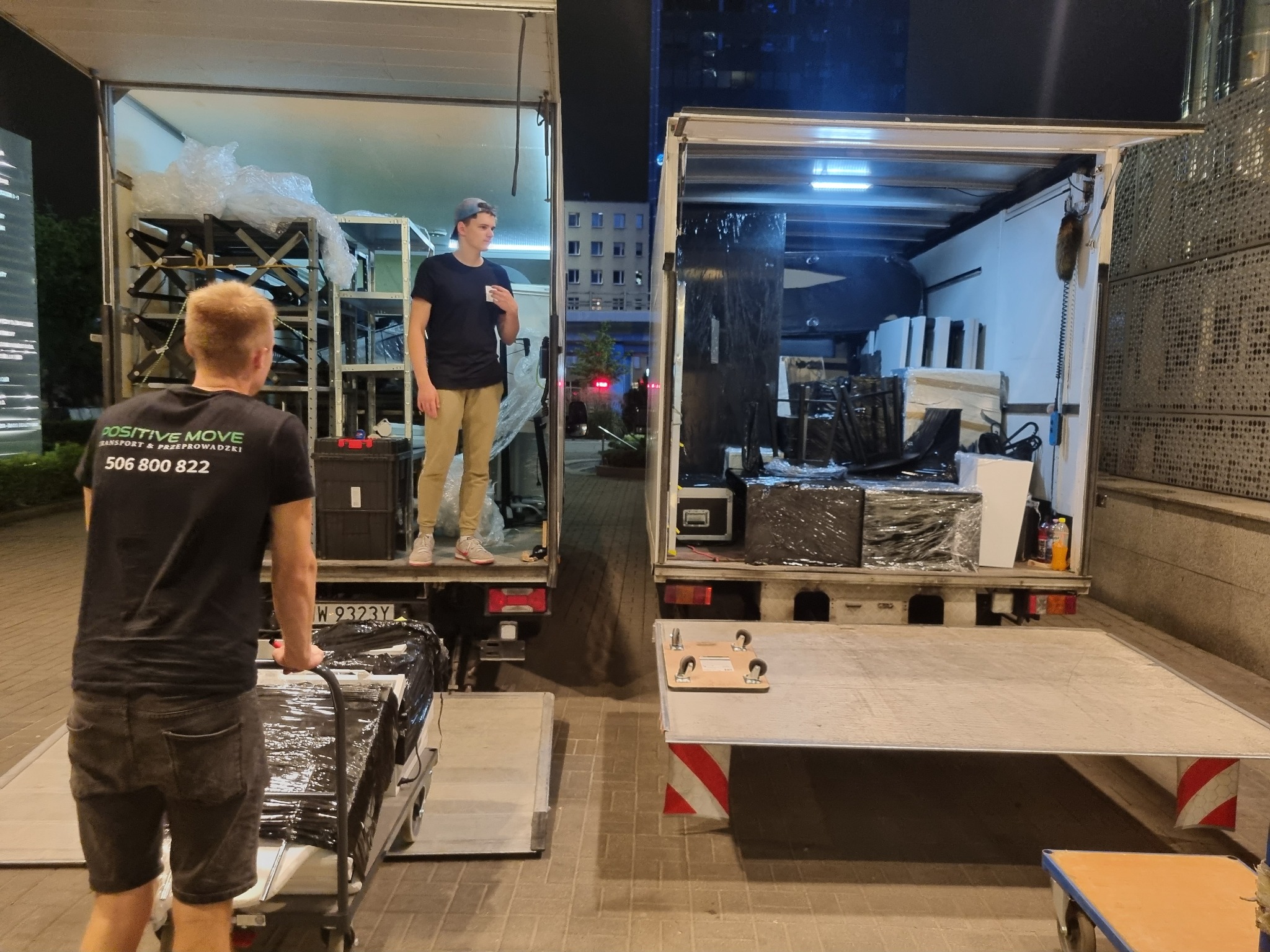 Dwa busy z otwartymi przestrzeniami ładunkowymi podczas załadunku, widoczni pracownicy w trakcie przenoszenia wyposażenia, jeden z wózkiem, drugi z kubkiem w ręku.