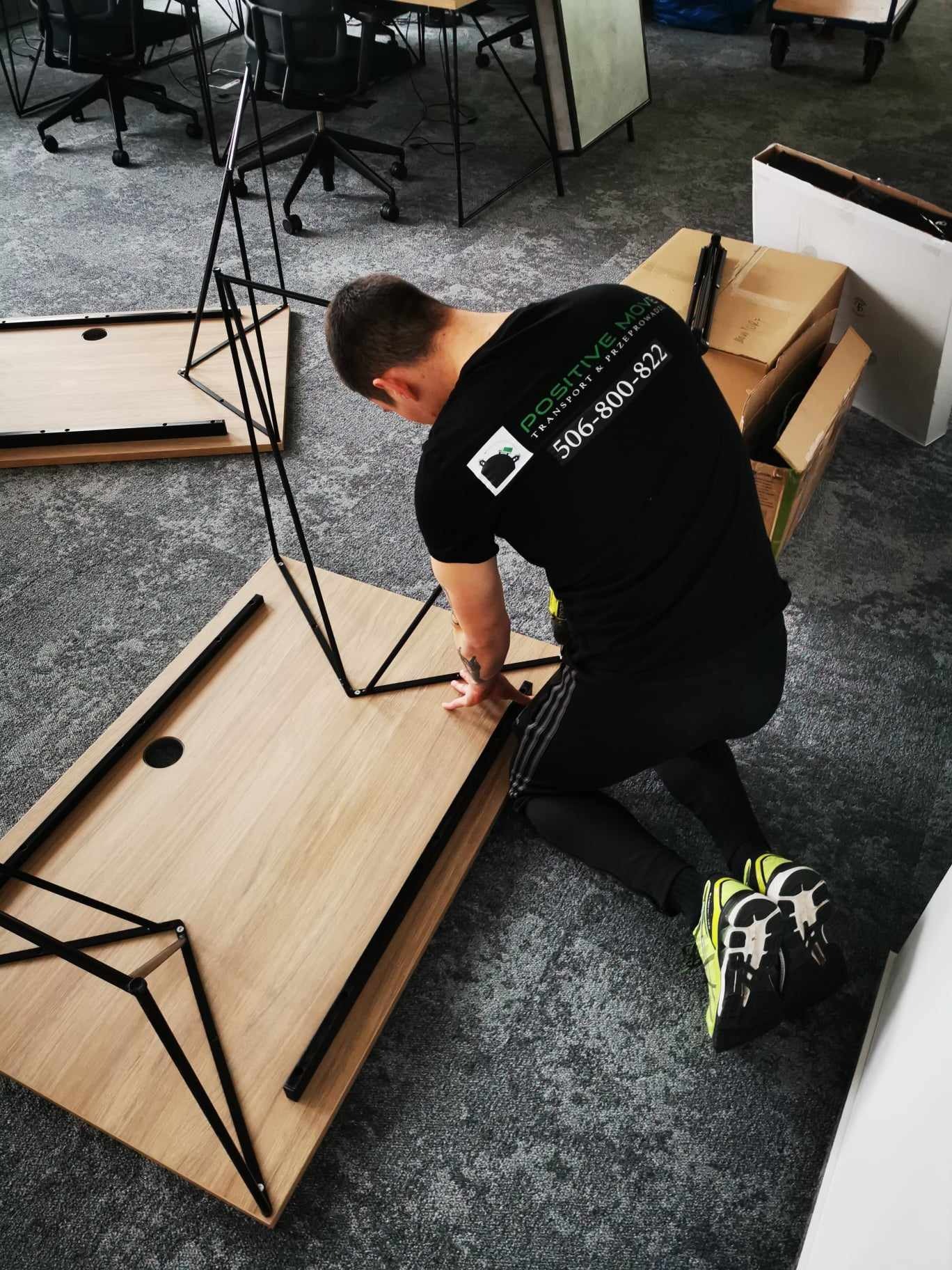 Pracownik firmy przeprowadzkowej montuje biurko w biurze, widoczne kartony i inne zdemontowane biurka w tle.