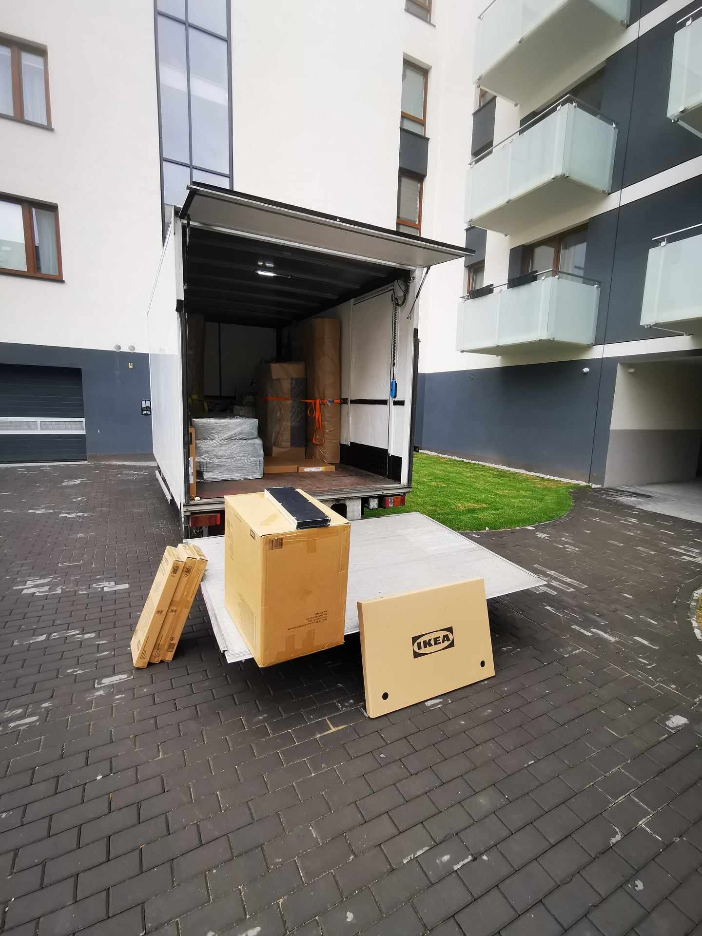Otwarty furgon transportowy załadowany meblami, z kartonami IKEA na rampie przed nowoczesnym budynkiem mieszkalnym.
