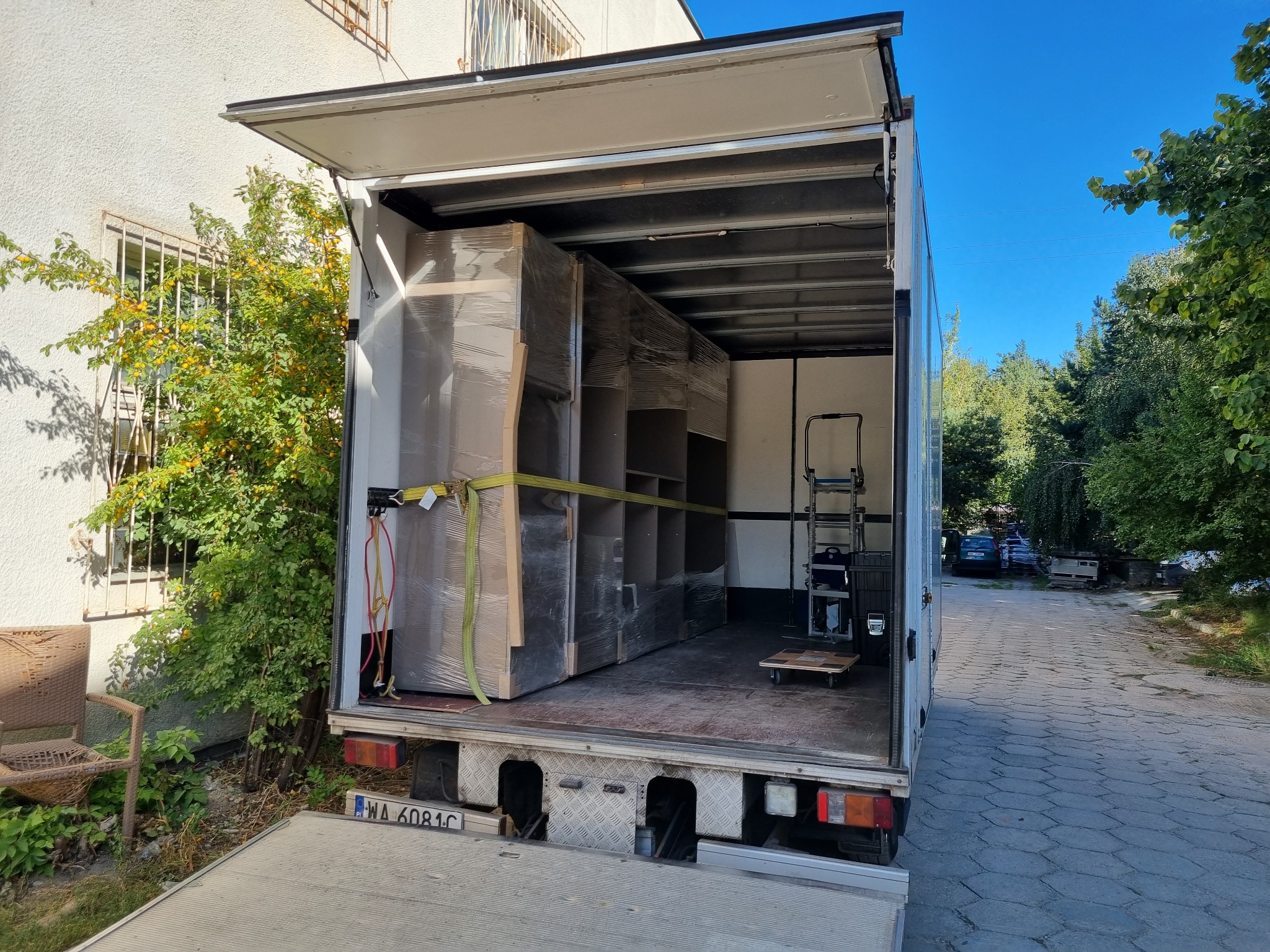 Wnętrze otwartego busa transportowego z załadowanymi, owiniętymi folią meblami zabezpieczonymi pasami, widoczna platforma załadunkowa i wózek transportowy, obok budynku z ozdobną kratą okienną.