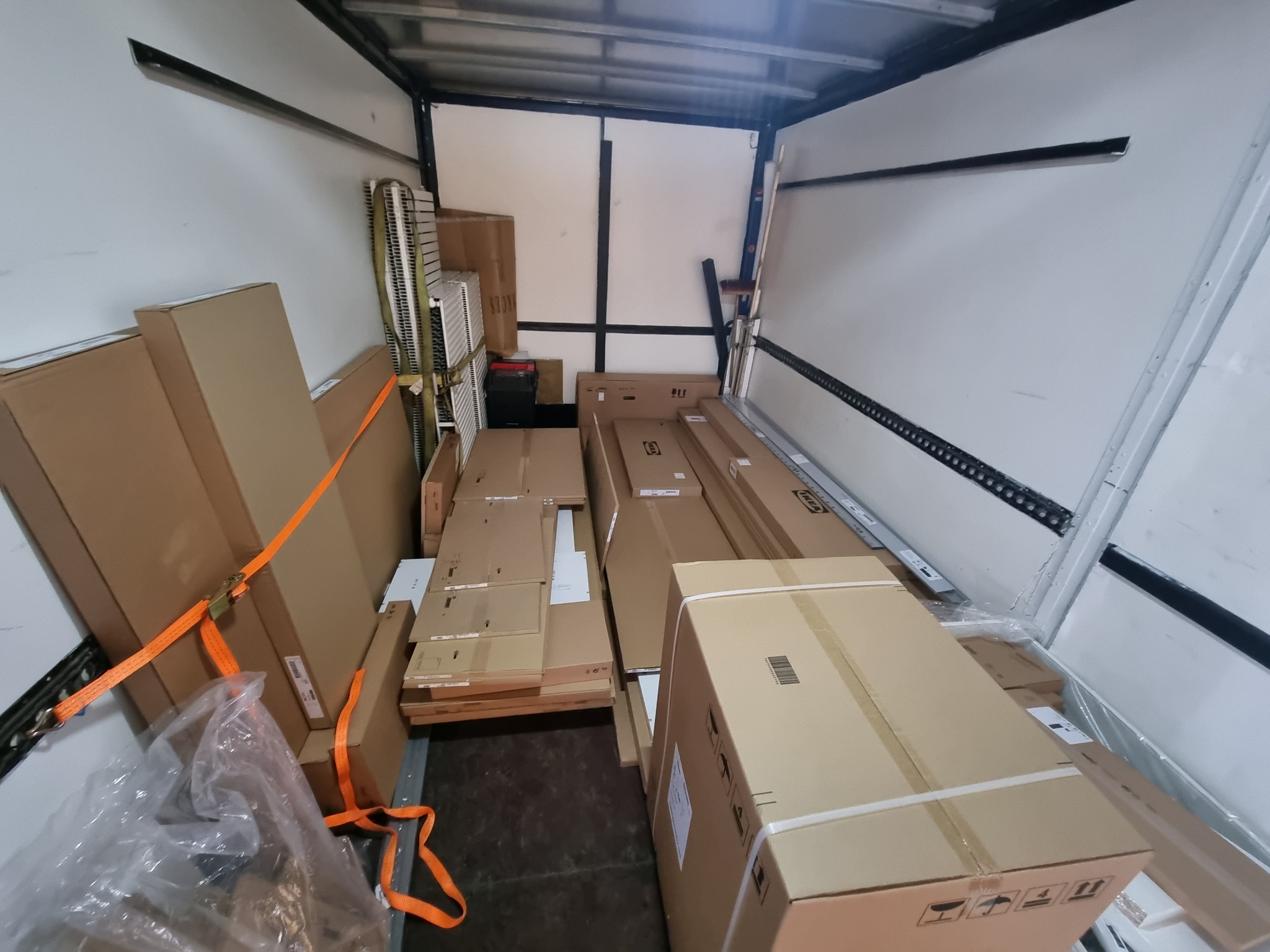 Wnętrze furgonetki wypełnionej kartonami, zabezpieczonymi pomarańczowymi pasami transportowymi, niektóre z logo IKEA, gotowe do transportu.