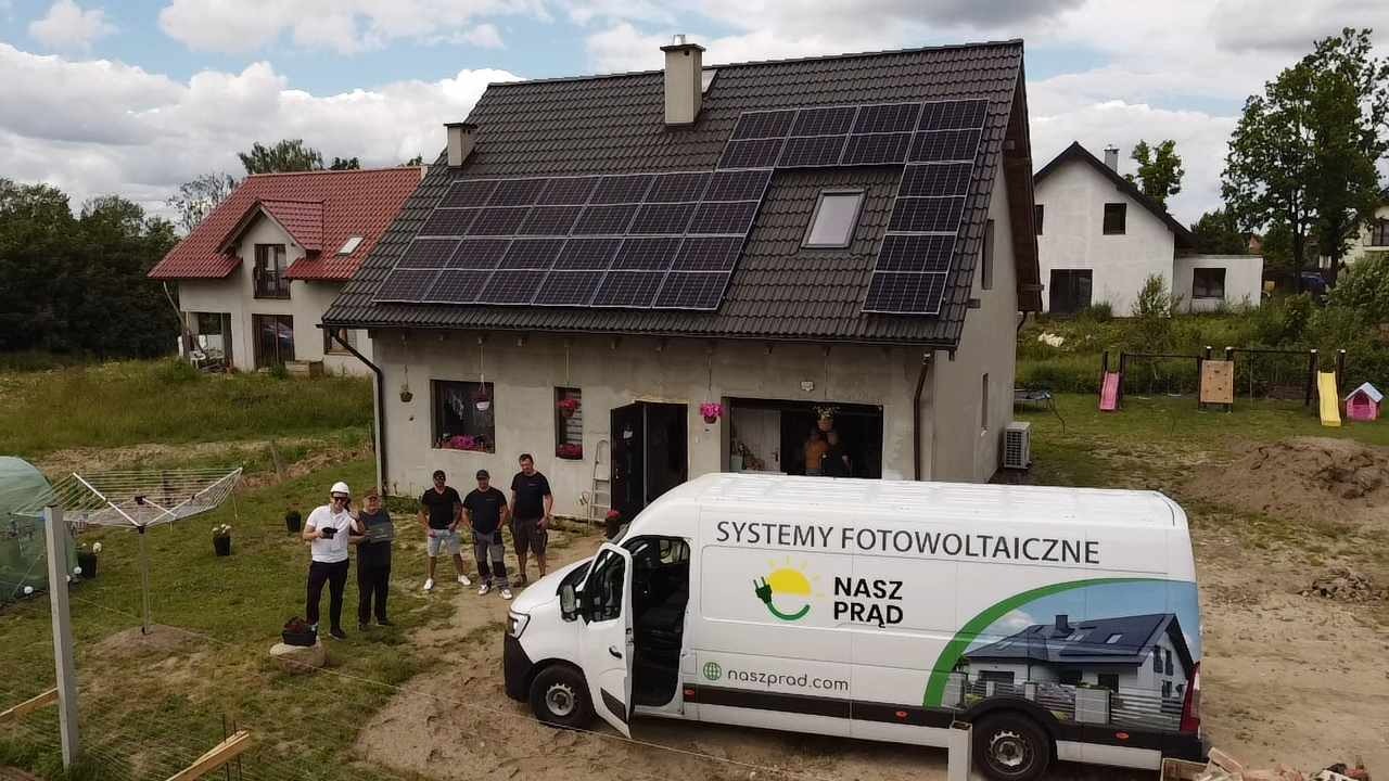 Dom jednorodzinny z szarą dachówką i panelami słonecznymi na dachu, ekipa montażowa oraz samochód firmowy z logo i hasłem 'Systemy Fotowoltaiczne Nasz Prąd'.
