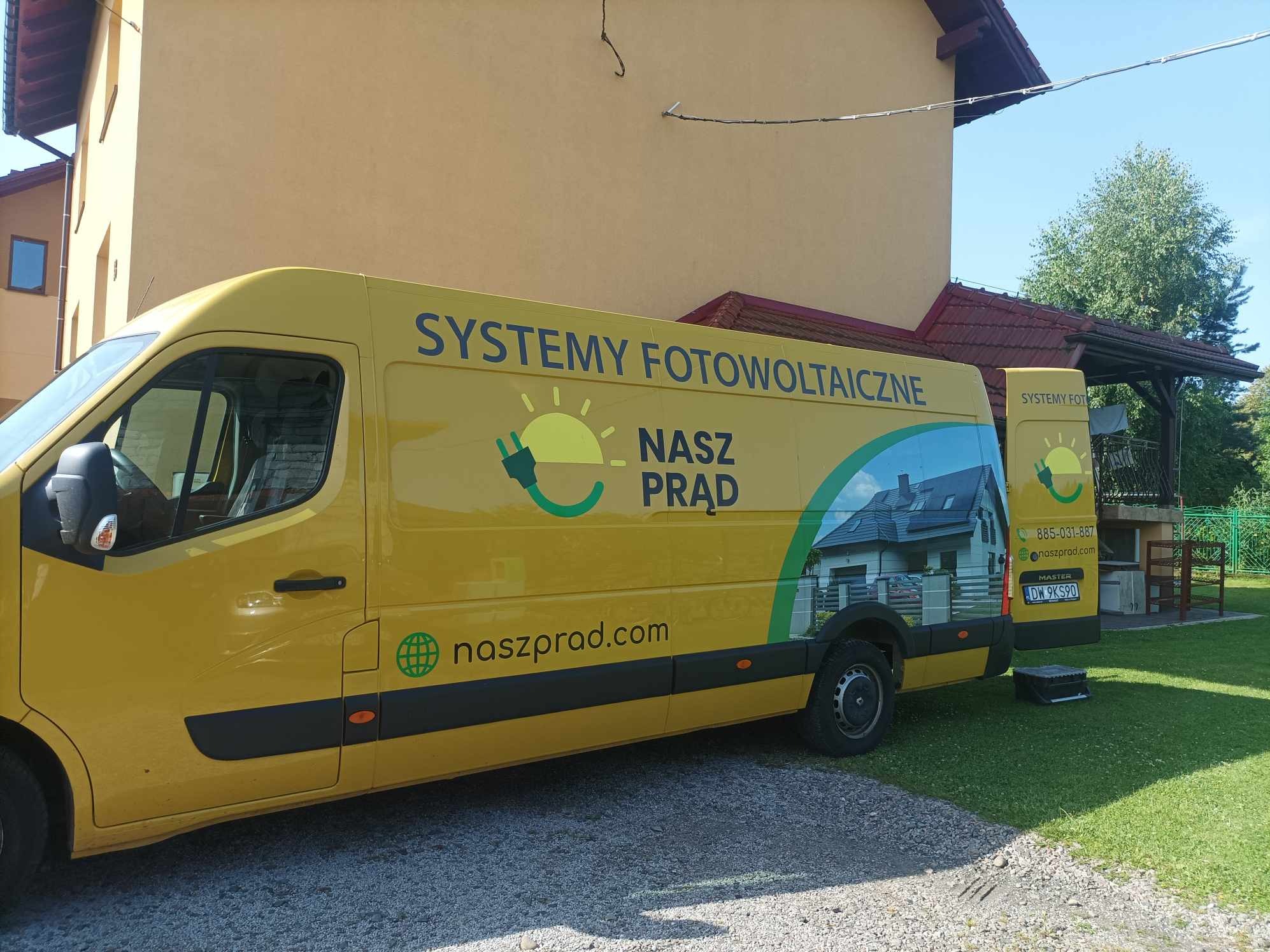 Żółty samochód dostawczy z grafiką przedstawiającą instalację paneli słonecznych na dachu domu, zaparkowany na żwirowym podjeździe przed budynkiem.