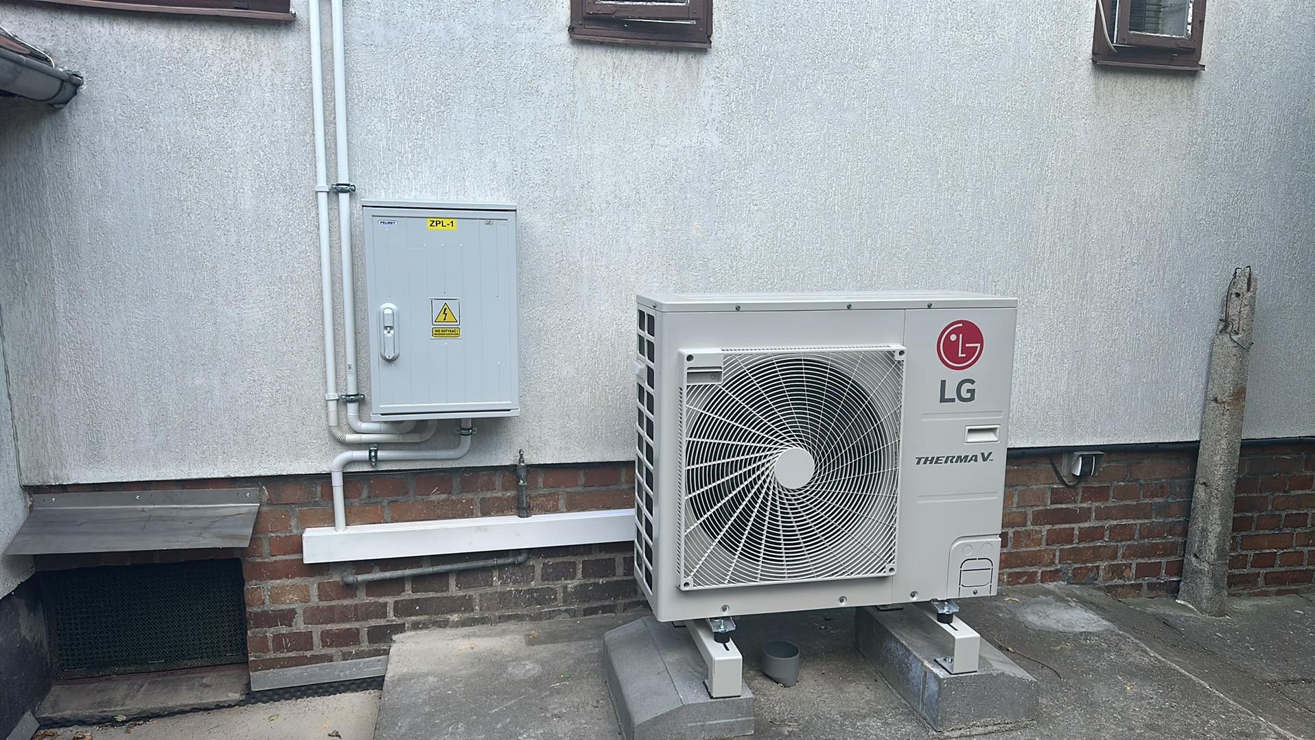 Zewnętrzna jednostka pompy ciepła LG Therma V zamontowana na betonowym fundamencie, obok skrzynka elektryczna ZPL-1, widoczne rury instalacyjne na tle otynkowanej ściany budynku.