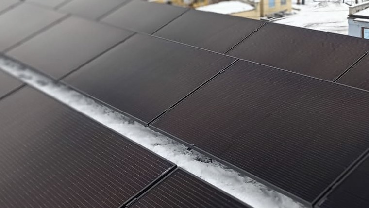 Częściowo ośnieżone, czarne panele słoneczne na dachu budynku w zimowy dzień.