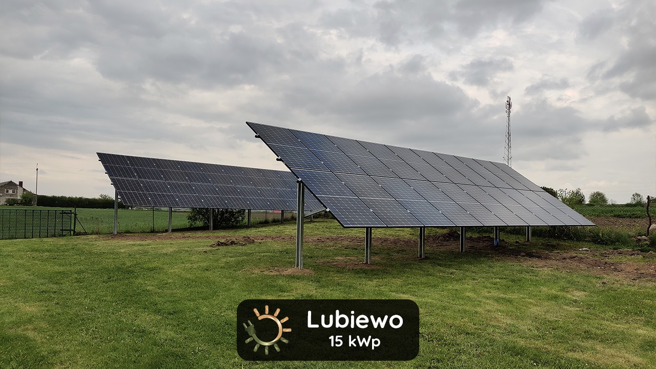 Dwa rzędy paneli słonecznych na metalowych podporach, na trawiastym terenie z widoczną wieżą telekomunikacyjną w tle.