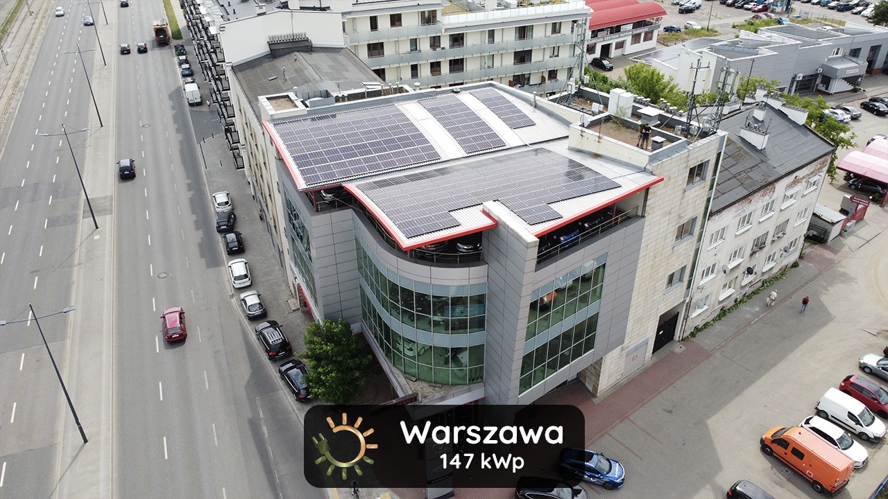 Widok z góry na budynek w Warszawie z panelami fotowoltaicznymi na dachu, z widoczną ulicą i zaparkowanymi samochodami.