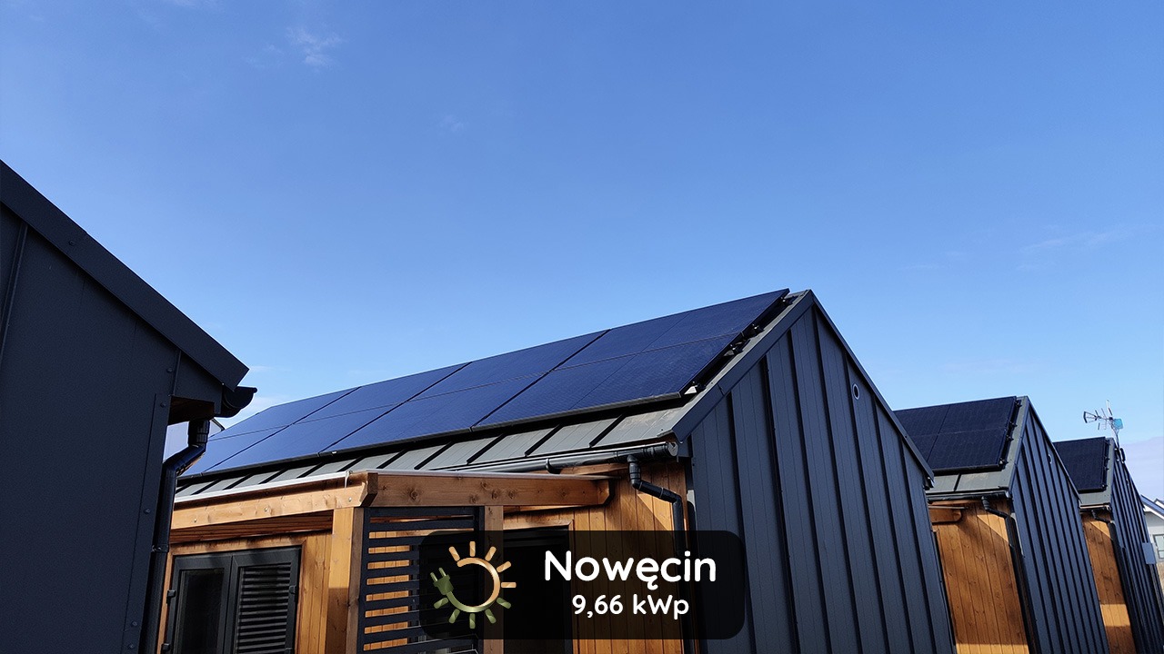 Panele słoneczne na dachu nowoczesnego domu modułowego z drewnianą elewacją i grafitową blachą trapezową, błękitne niebo w tle.