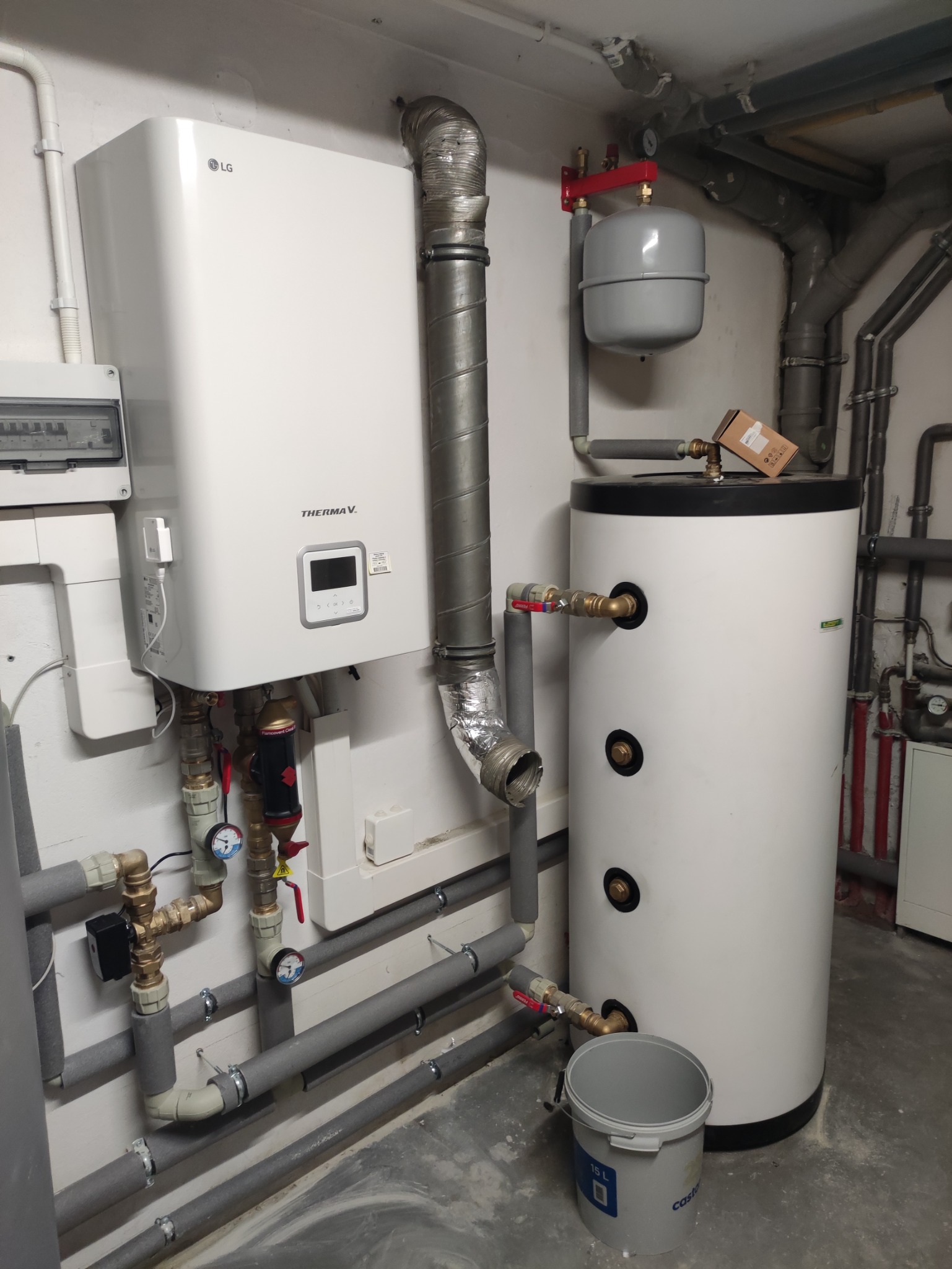 Biała pompa ciepła LG Therma V z podłączonymi rurami i zbiornikiem na wodę, widoczny również szary kubeł na podłodze.