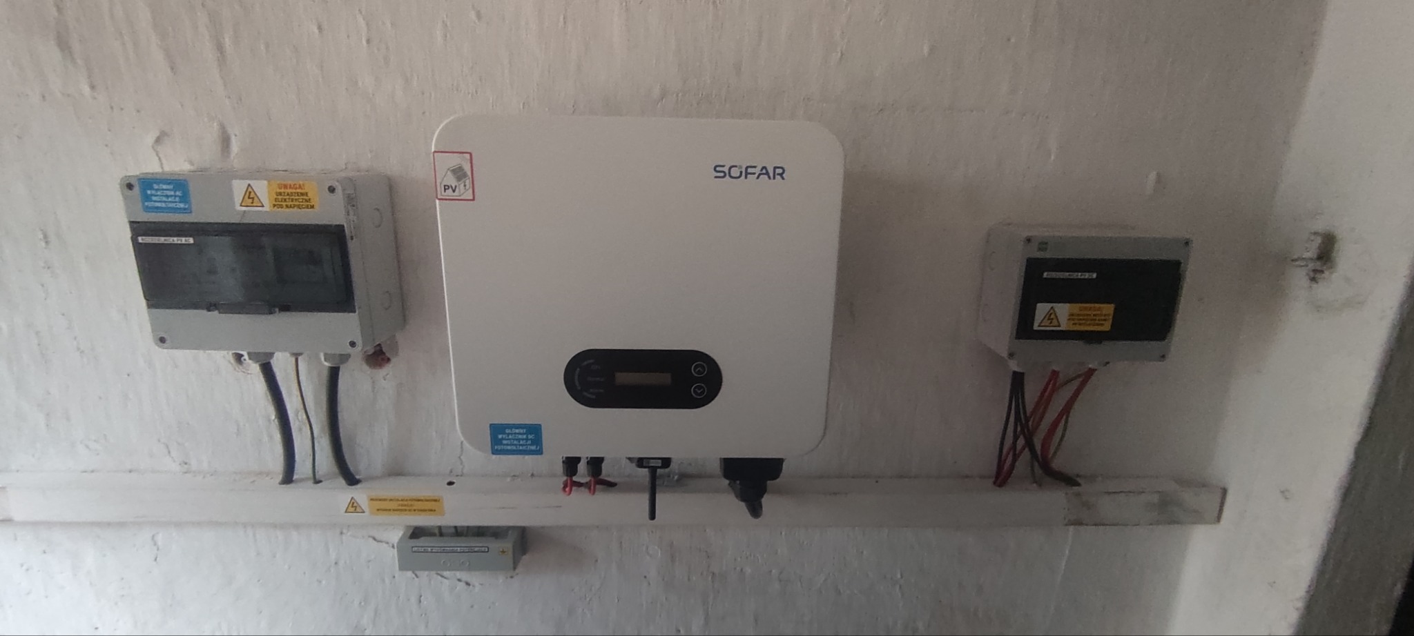 Zamontowany inwerter SOFAR z dwoma skrzynkami elektrycznymi na pomalowanej ścianie o nierównej fakturze, widoczne kable i naklejki ostrzegawcze.