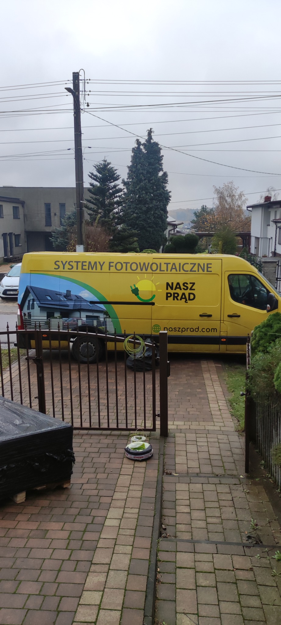 Żółty samochód dostawczy z napisem 'Systemy Fotowoltaiczne' i grafiką domu z panelami słonecznymi, zaparkowany na brukowanej posesji.