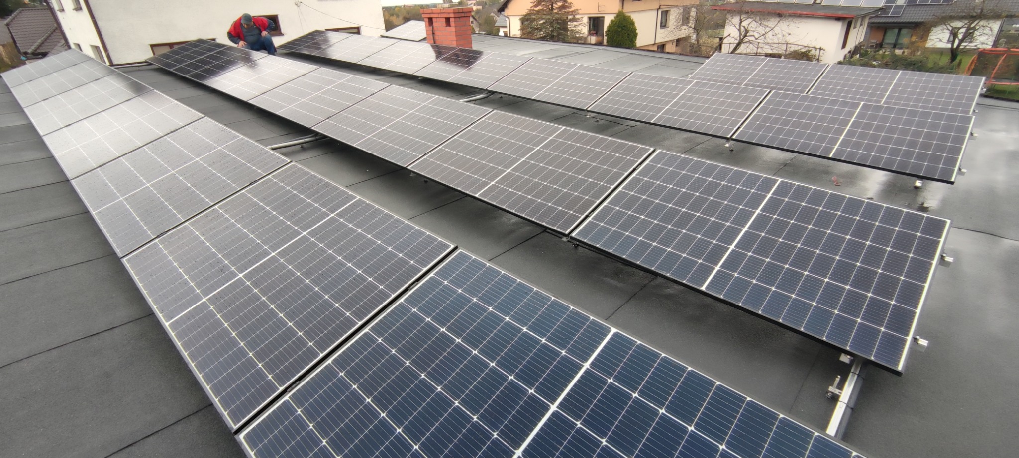 Montaż paneli słonecznych na płaskim dachu, widoczny pracownik w czerwonej kurtce, w tle zabudowa jednorodzinna.