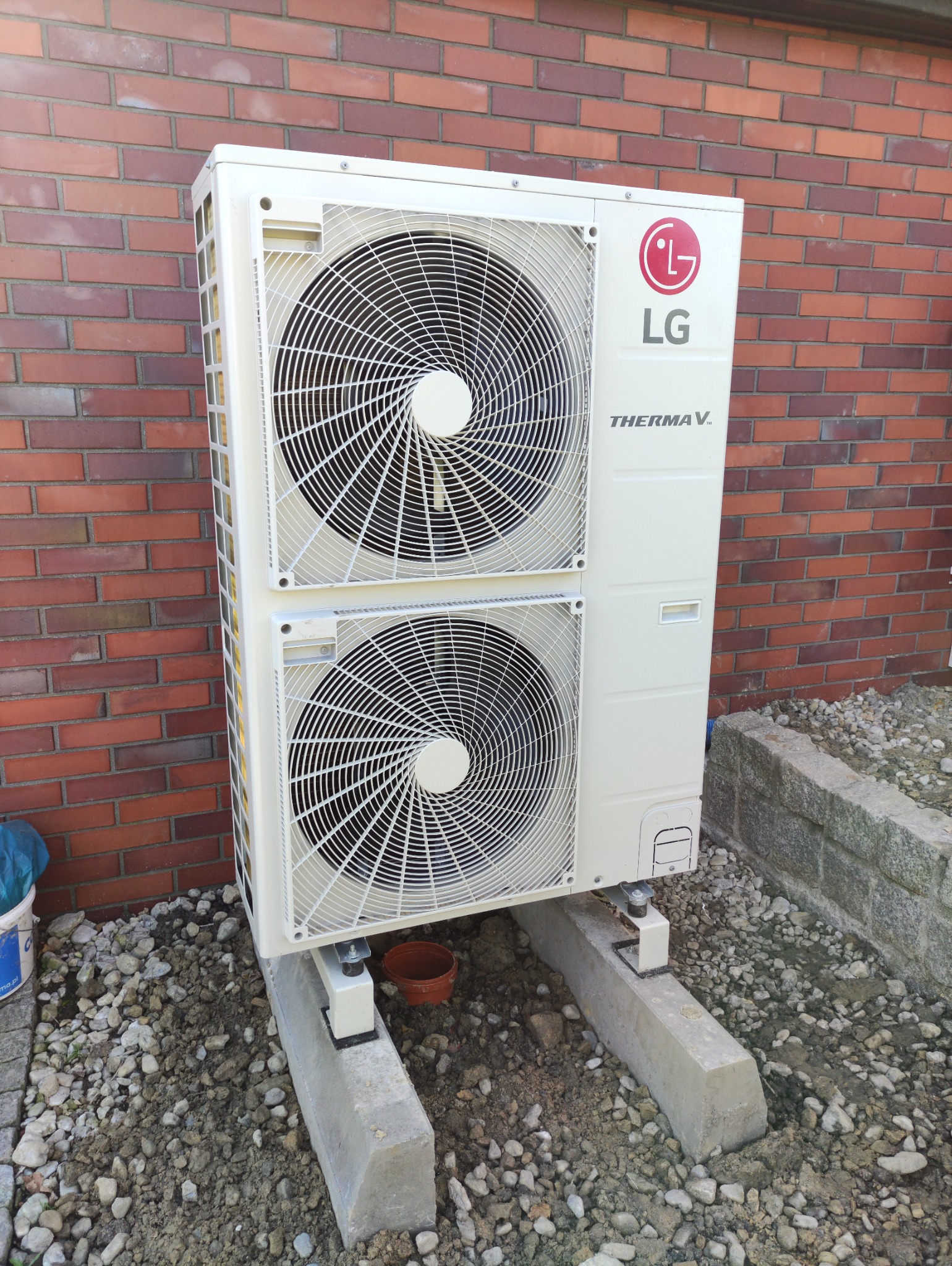 Biała, dwusekcyjna zewnętrzna jednostka pompy ciepła LG Therma V, zainstalowana na betonowych podstawach obok ceglanego budynku, widoczne dwa wentylatory i logo LG.