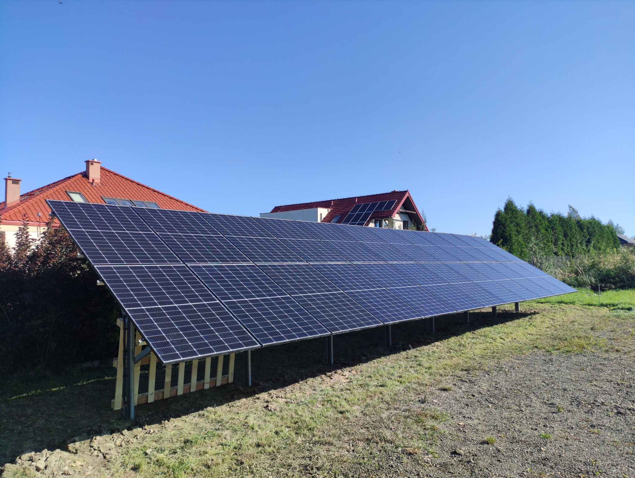 Panele słoneczne ustawione na ziemi przed domem z czerwoną dachówką pod błękitnym niebem.