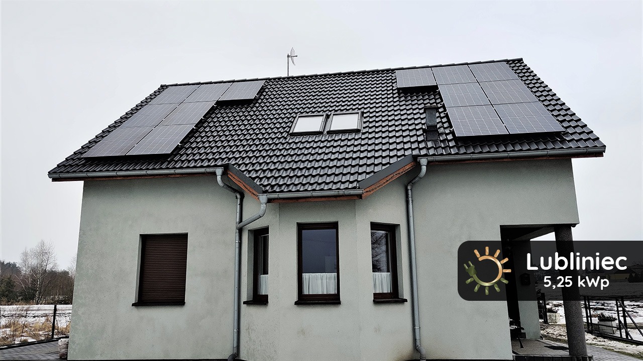 Dom jednorodzinny z panelami słonecznymi na dachu pokrytym czarną dachówką, widoczne okna dachowe i antena, rynny odprowadzające wodę, na elewacji budynku okna z roletami i zasłonami, w rogu kadru...