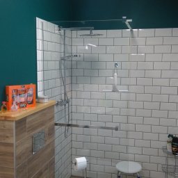 INTERIOR - Nowoczesna kabina prysznicowa z białymi, prostokątnymi płytkami, szklaną ścianką i deszczownicą, uzupełniona o drewnopodobną zabudowę z zestawem kosmetyków i rośliną doniczkową, na tle ściany...