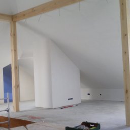 INTERIOR - Poddasze w trakcie prac wykończeniowych: widoczne drewniane belki konstrukcyjne, białe ściany, okno dachowe, rusztowanie z zielonym ręcznikiem, skrzynka z narzędziami i kabel na podłodze.