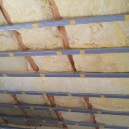 INTERIOR - Poddasze z izolacją z wełny mineralnej, widoczne drewniane krokwie i metalowe profile mocujące, zabezpieczone żółtą taśmą.