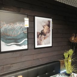 INTERIOR - Ciemna drewniana ściana z dwoma obrazami w czarnych ramach, lampą wiszącą z miedzianymi kloszami i zieloną dekoracyjną trawą w doniczce, widoczny fragment wezgłowia łóżka.