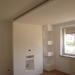 INTERIOR - Wnętrze pokoju w trakcie prac: zabudowa z płyty gipsowo-kartonowej z wnękami na sprzęt RTV i półkami narożnymi, widoczne otwory na przewody, surowa podłoga, okno z żaluzjami.