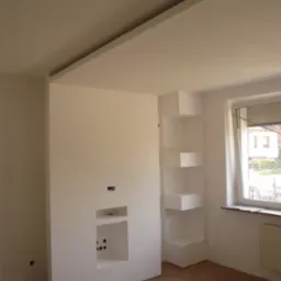 Wnętrze pokoju w trakcie prac: zabudowa z płyty gipsowo-kartonowej z wnękami na sprzęt RTV i półkami narożnymi, widoczne otwory na przewody, surowa podłoga, okno z żaluzjami.