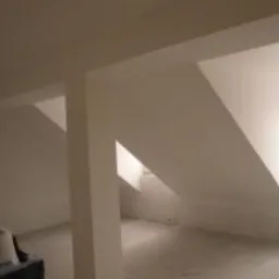 Białe, wykończone poddasze z widocznymi skośnymi ścianami, kolumną nośną i oknem dachowym, w trakcie prac wykończeniowych.