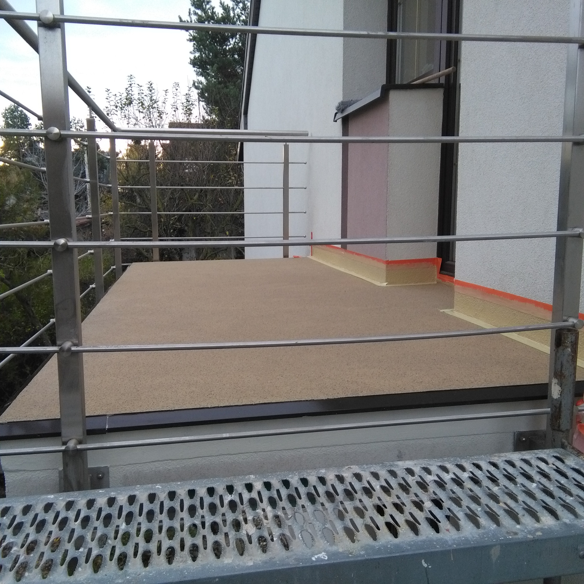 Balkon z nową, beżową, żywiczną posadzką i metalową balustradą, widok z zewnątrz, z bliska widoczna taśma malarska zabezpieczająca krawędzie przy ścianie budynku.