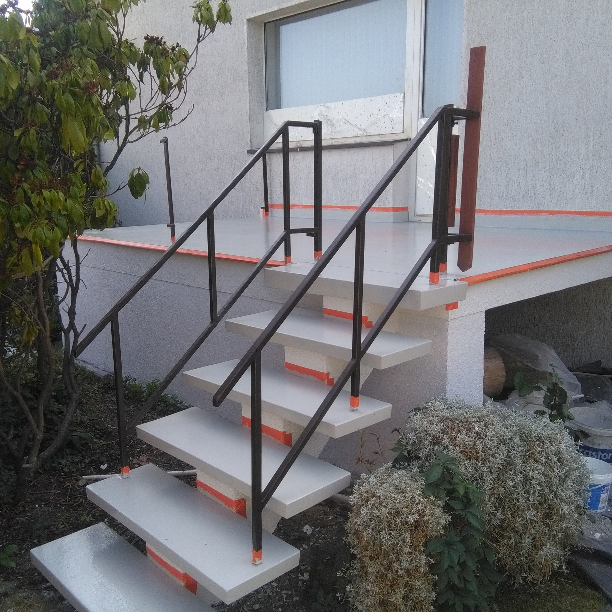 Zewnętrzne schody betonowe z czarną metalową balustradą, wykończone pomarańczowymi akcentami, prowadzące do wejścia domu z widocznym oknem i roślinnością w tle.